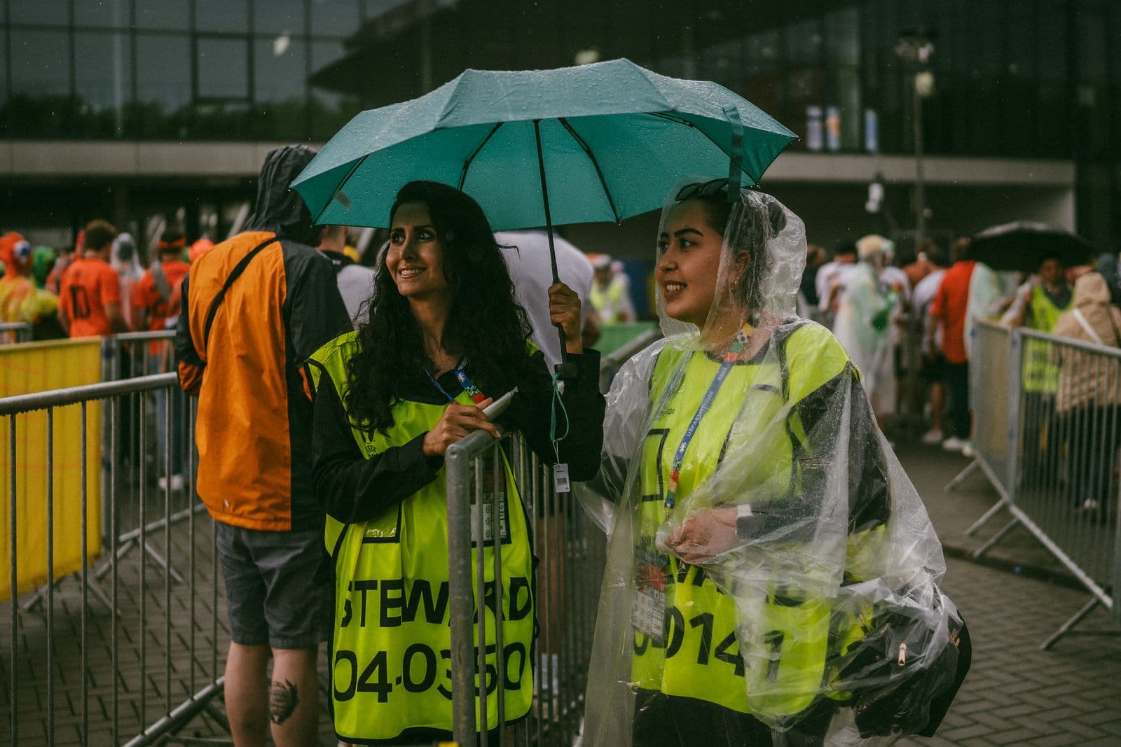Two smiling event stewards in high-visibility vests sharing an umbrella at a crowd barrier