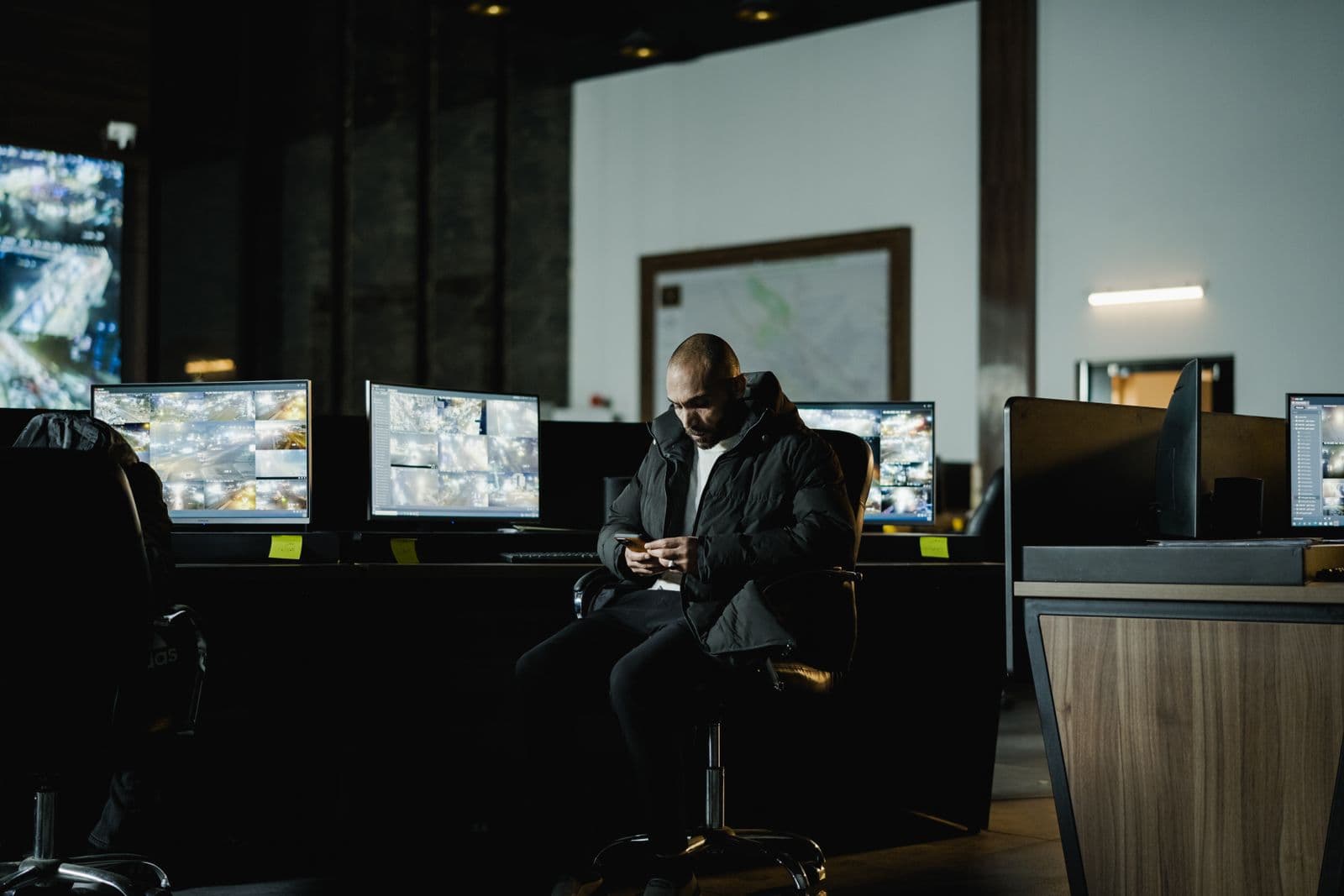 A security supervisor alone in a dark control room, checking his phone in front of camera monitors