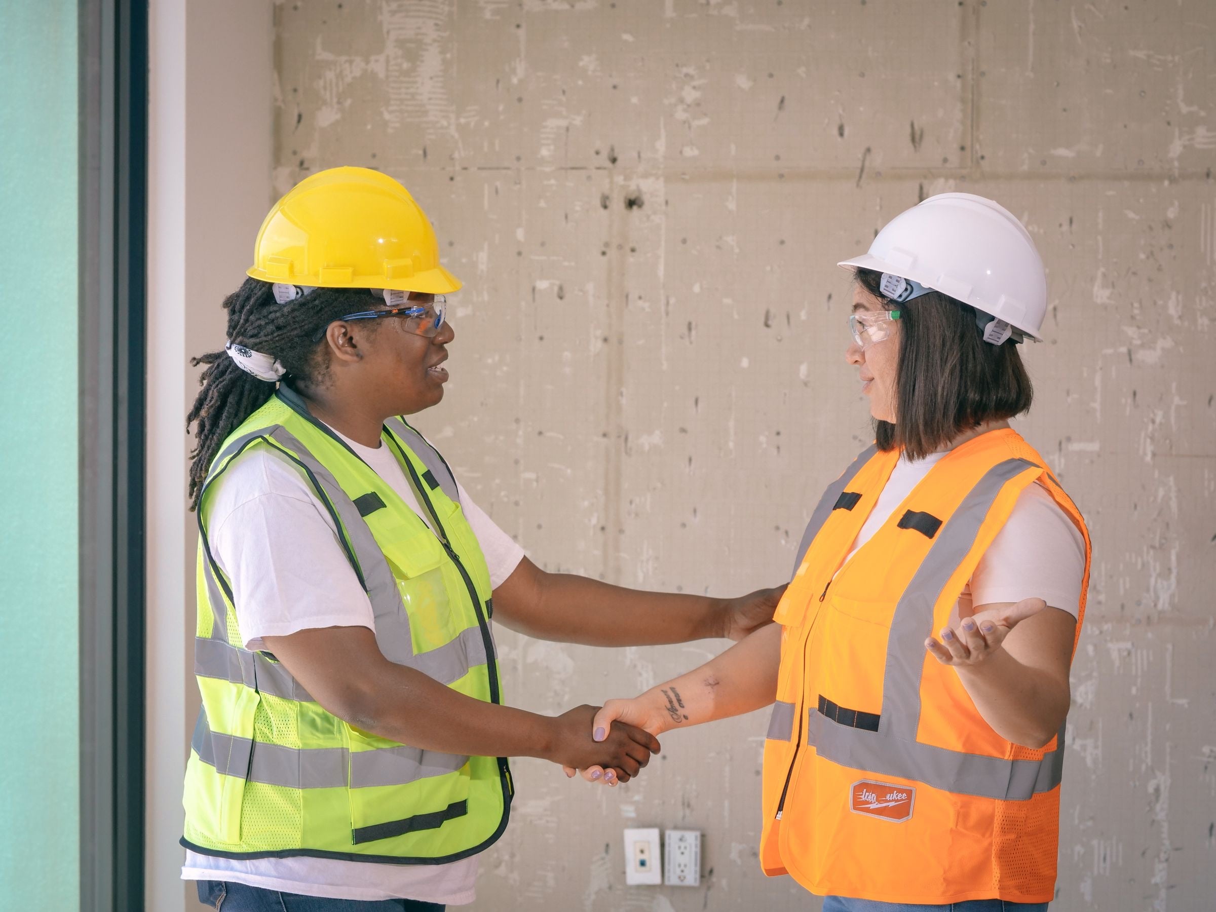 Two women in hard hats and hi-vis vests shake hands and smile