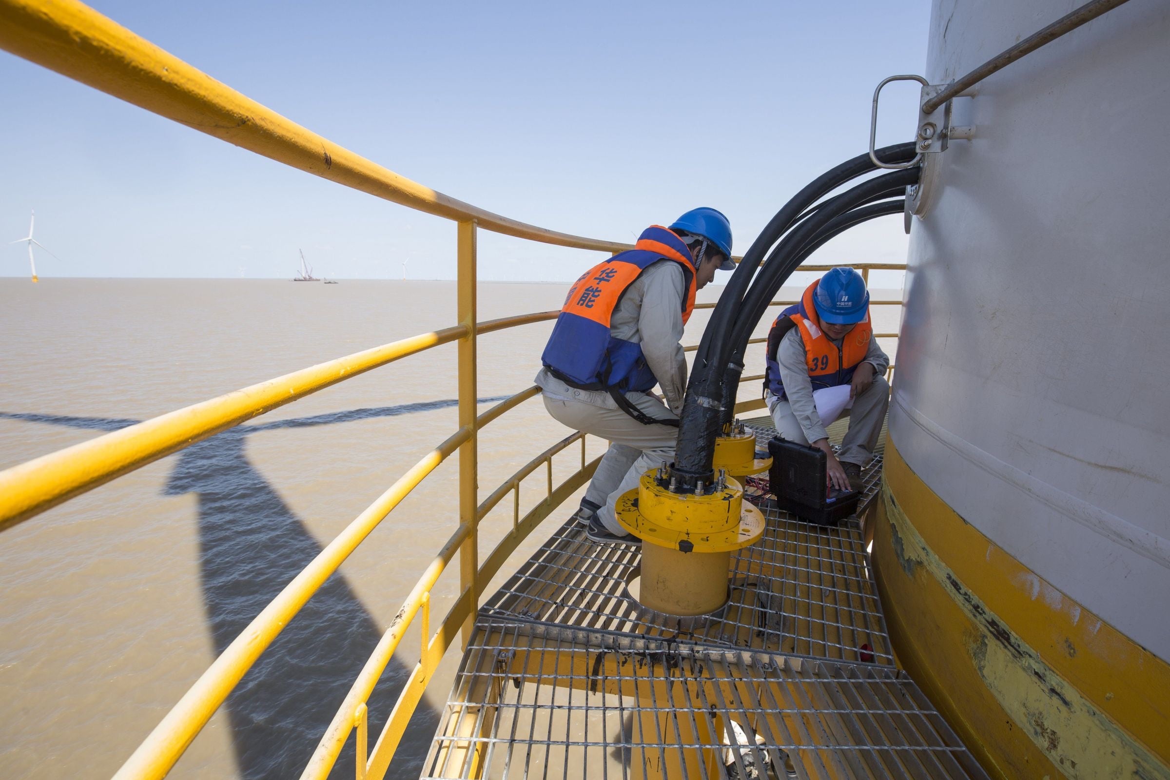 Two technicians in hard hats and life jackets work on the platform of an offshore wind turbine