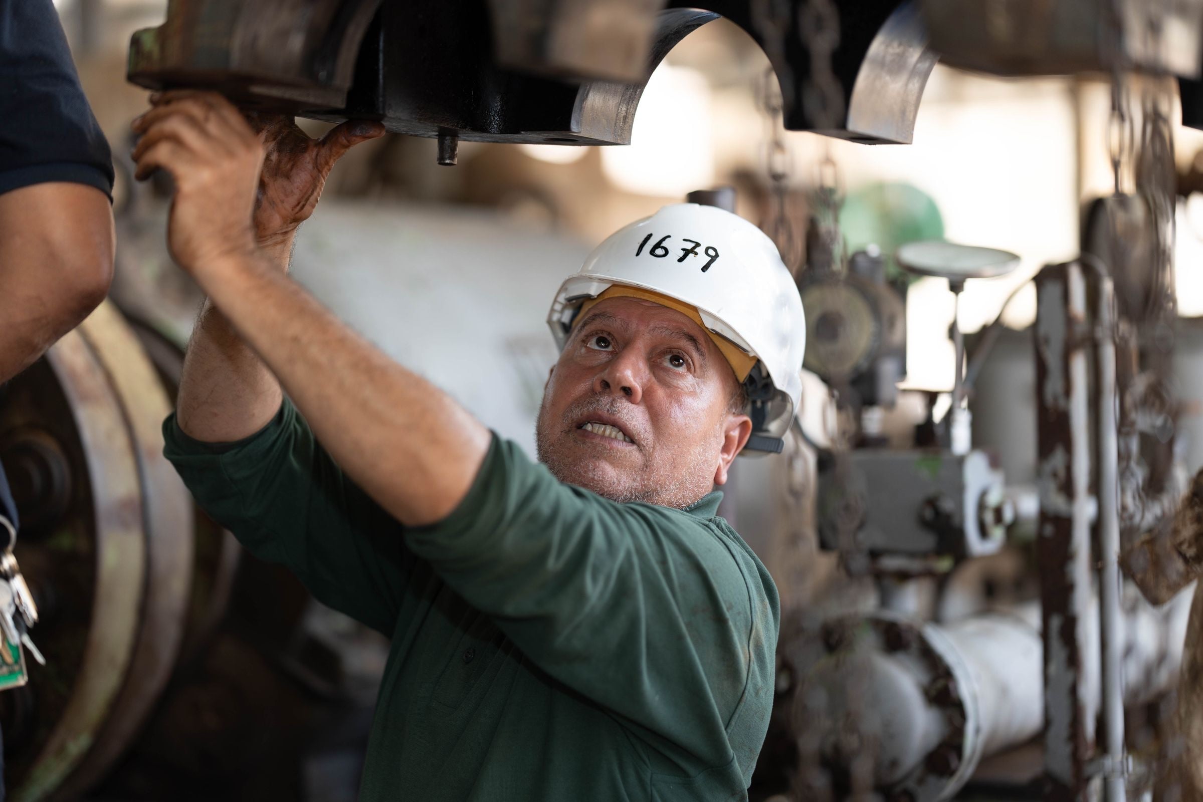 A maintenance fitter in a white hard hat reaches up to work on heavy plant machinery