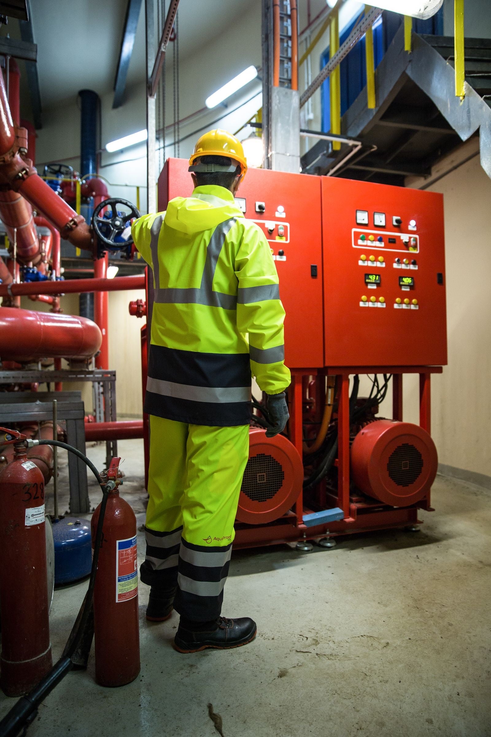 A worker in a hard hat and yellow hi-vis overalls checks a red pump control panel in a plant room