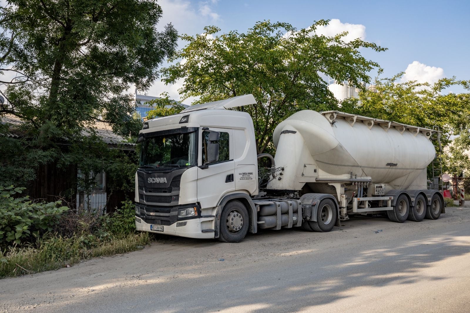 A white Scania tractor unit pulling a bulk powder tanker, parked on a tree-lined street