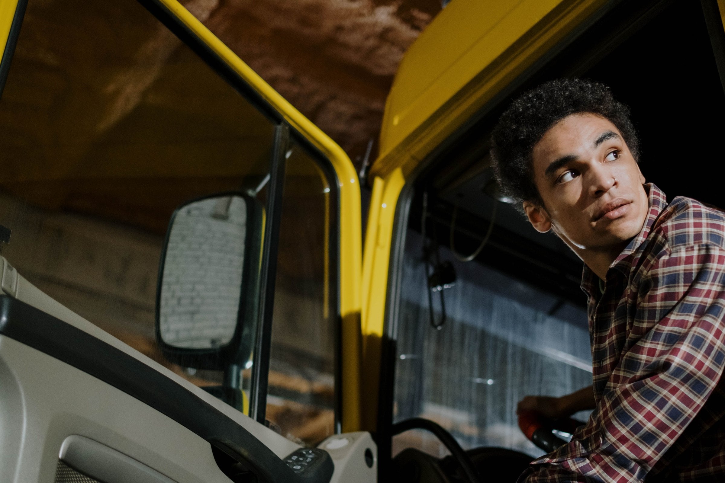 A young driver in a checked shirt sits in the open door of a yellow cab-over lorry, looking back over his shoulder