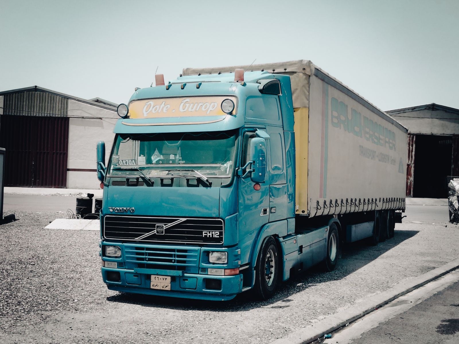 A teal Volvo tractor unit coupled to a curtainsider trailer, parked outside a warehouse