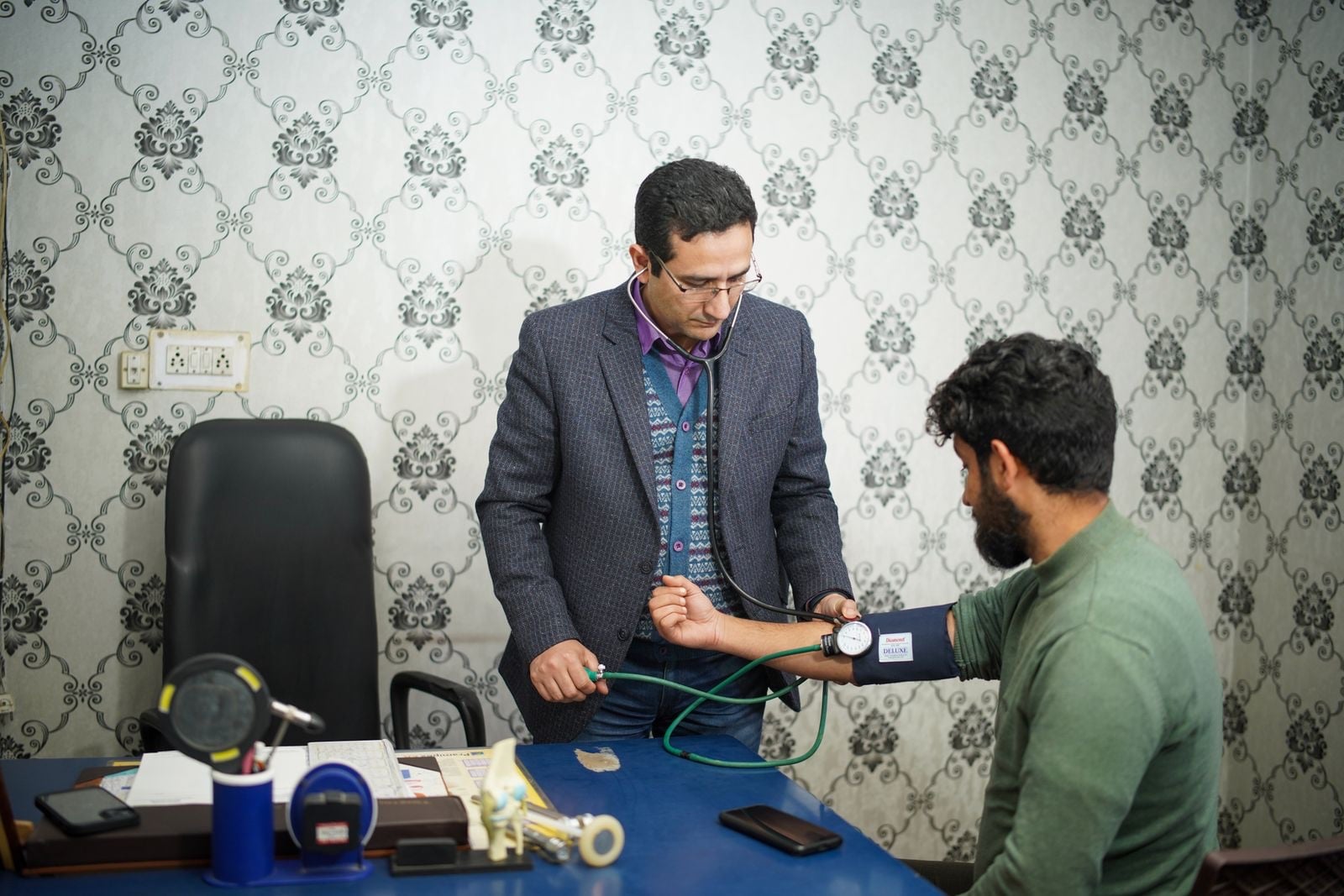 A doctor takes a bearded man’s blood pressure with a cuff at a desk in a consulting room