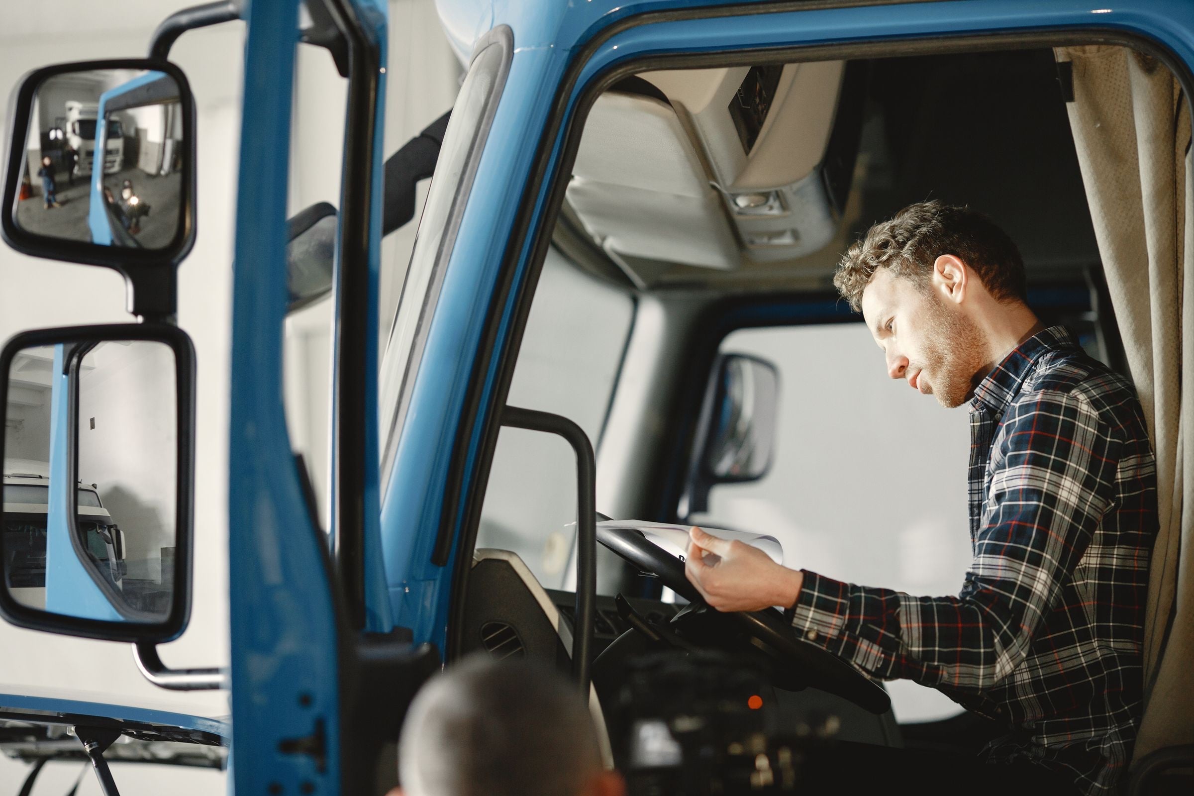 A driver in a checked shirt reads a sheet of paperwork in the cab of a blue lorry with the door open