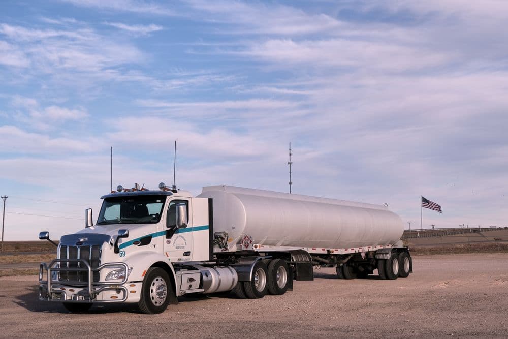 A white tanker truck parked on a gravel lot under a wide sky, a US flag in the distance