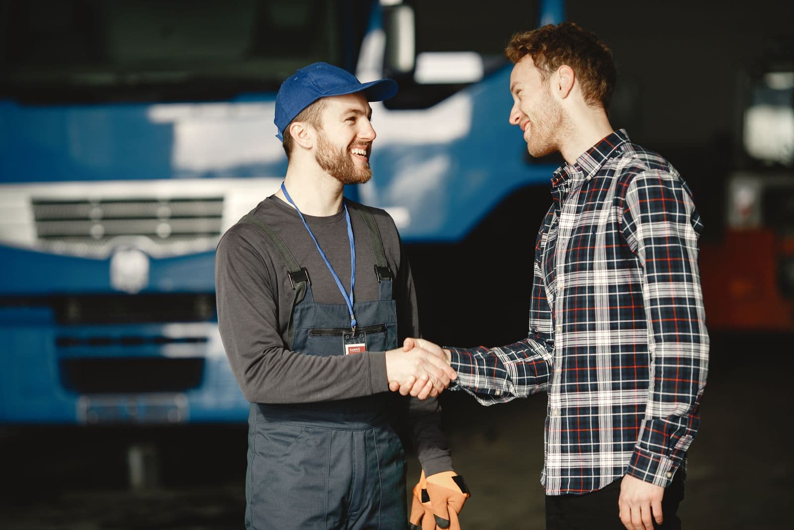 Two smiling men shake hands in front of a blue truck in a depot