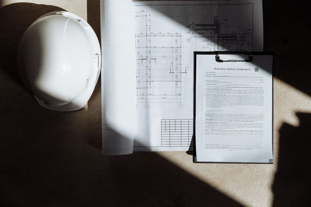 A white hard hat beside site plans and a clipboard of paperwork