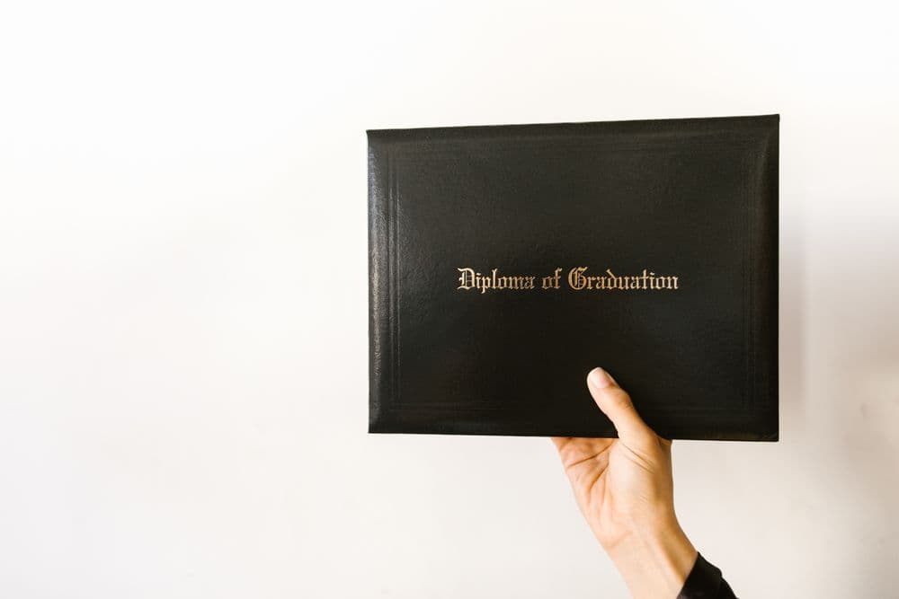 A hand holding up a black diploma cover against a white wall