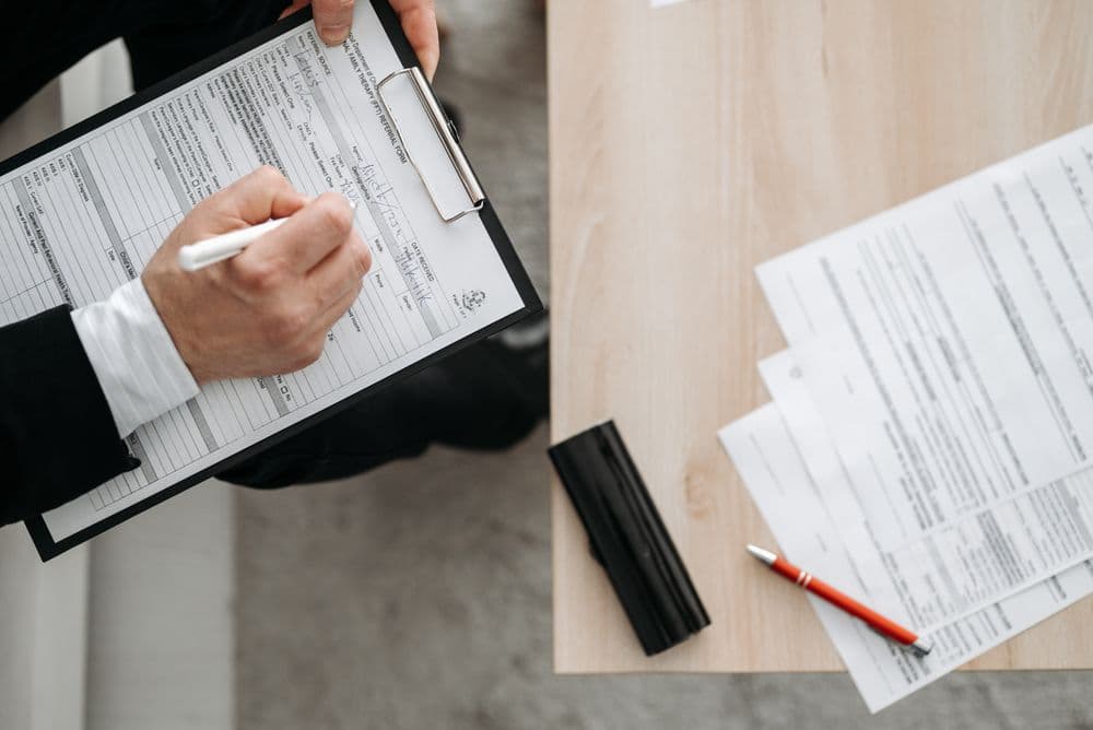 A person filling in a form on a clipboard beside a stack of paperwork