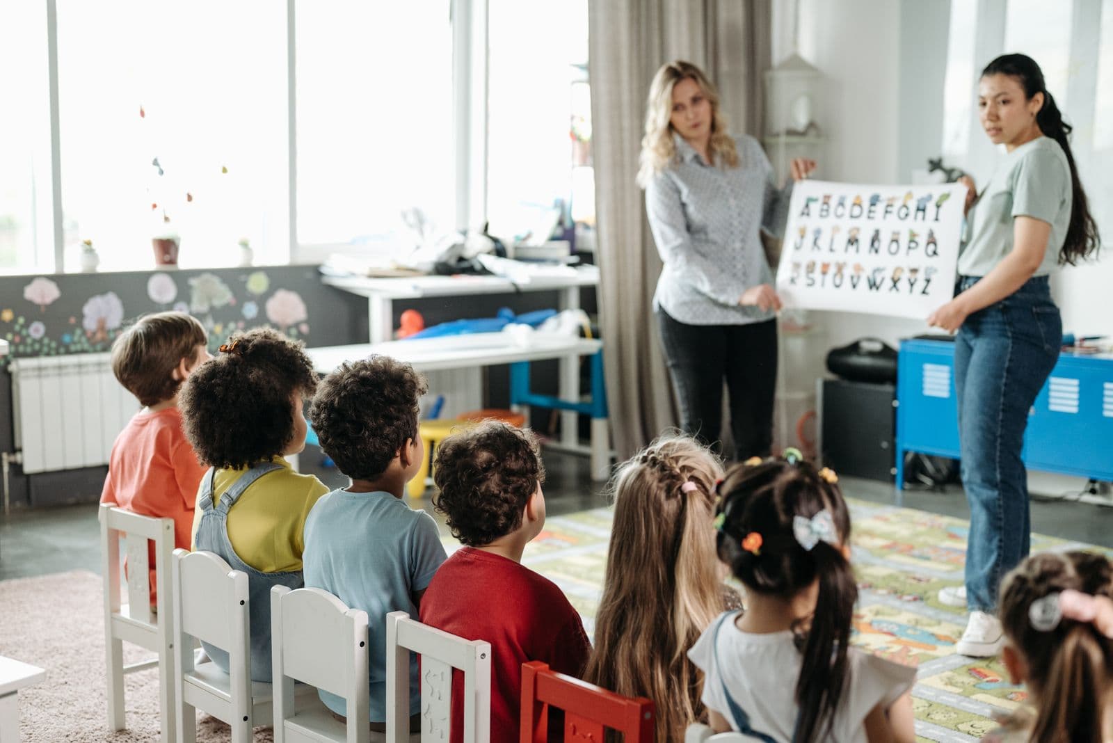 Two teachers holding up an alphabet chart for a kindergarten class seated on small chairs