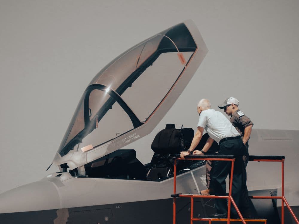 Two men on a red platform look into the open cockpit of a military jet