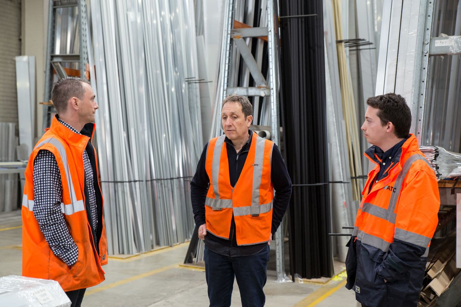 Three colleagues in high-visibility vests talking among racks of metal stock