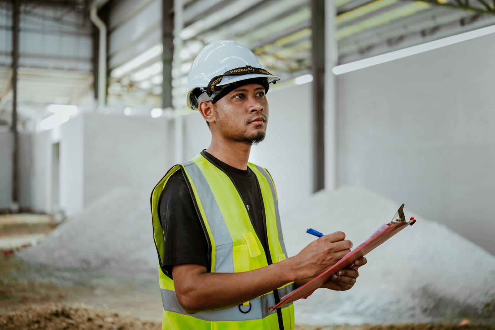 A supervisor in a hard hat and safety vest writes on a clipboard inside a plant