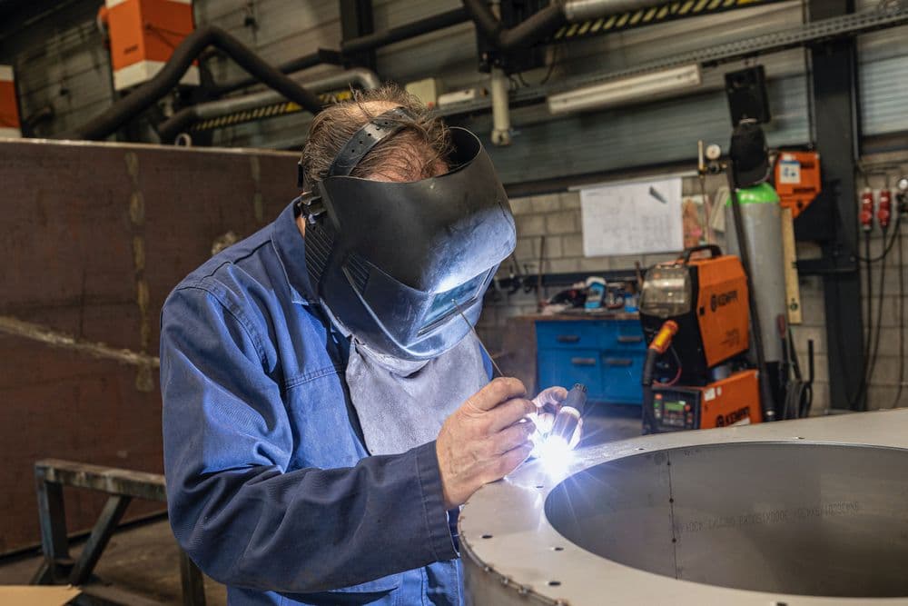 A welder in a welding helmet works on a steel ring in a fabrication shop