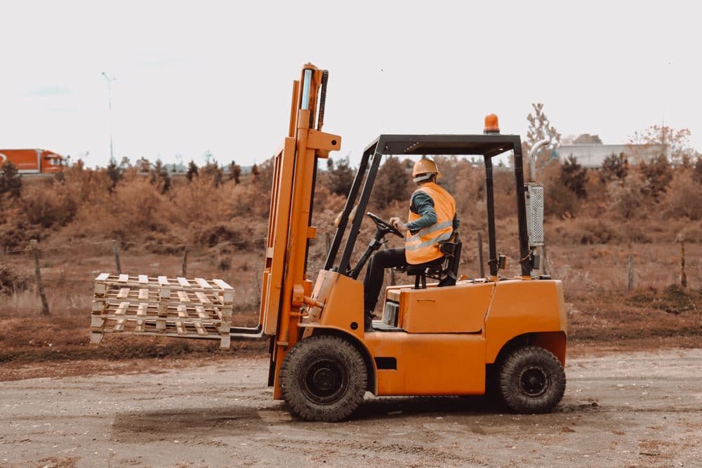 A worker in a hard hat and safety vest drives a forklift carrying empty pallets