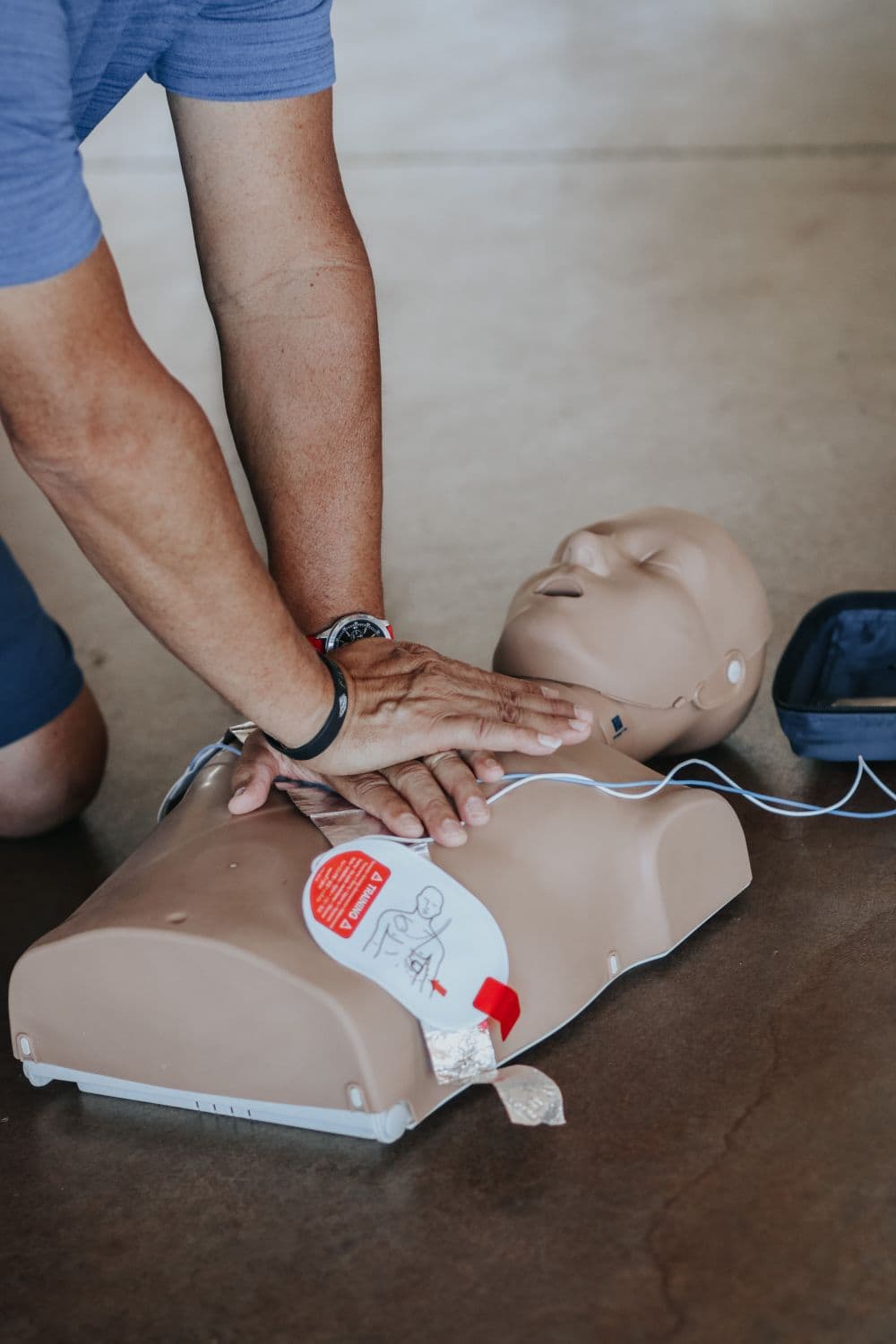 Hands performing CPR on a training manikin beside an AED