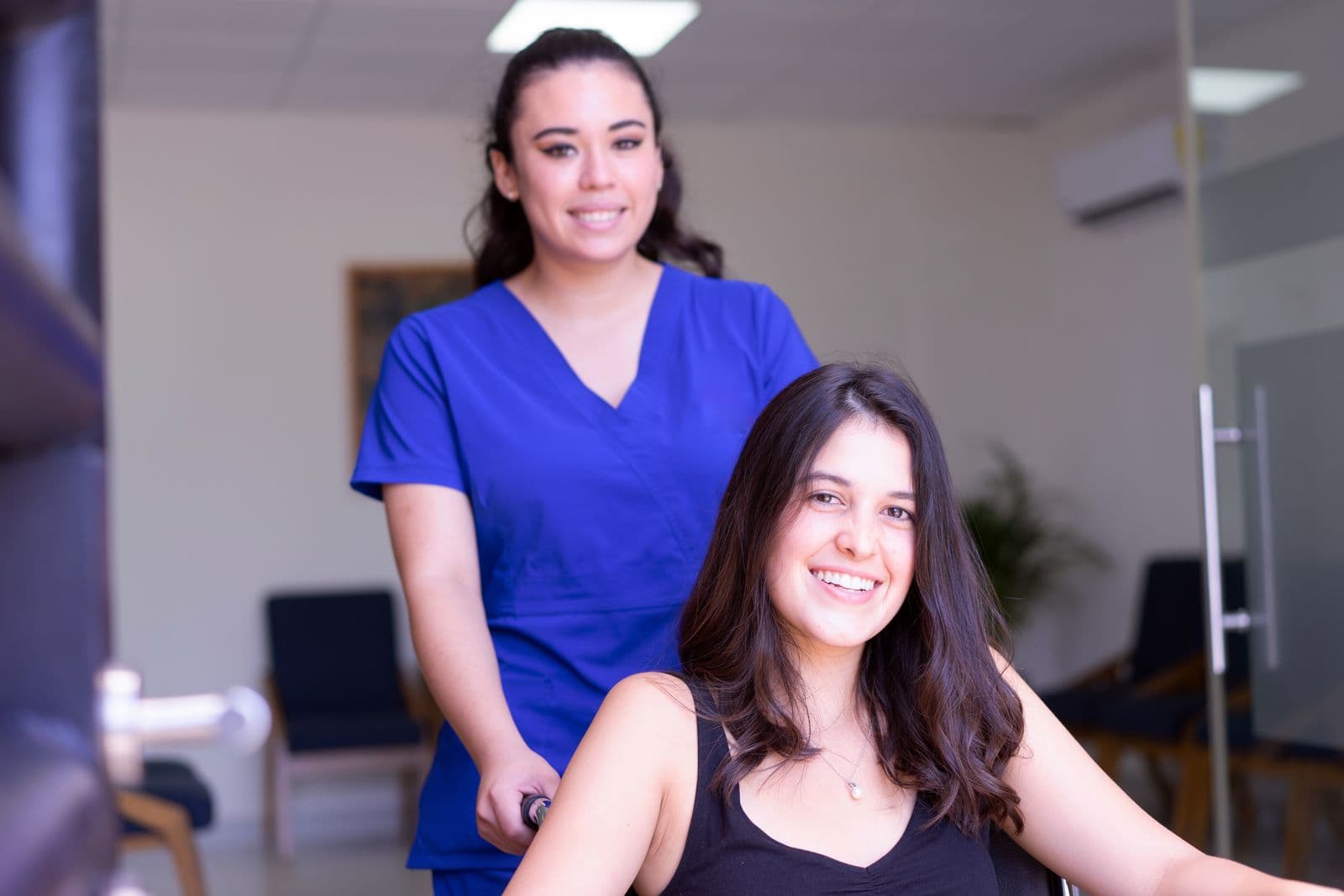 A nurse in blue scrubs with a smiling patient in a wheelchair