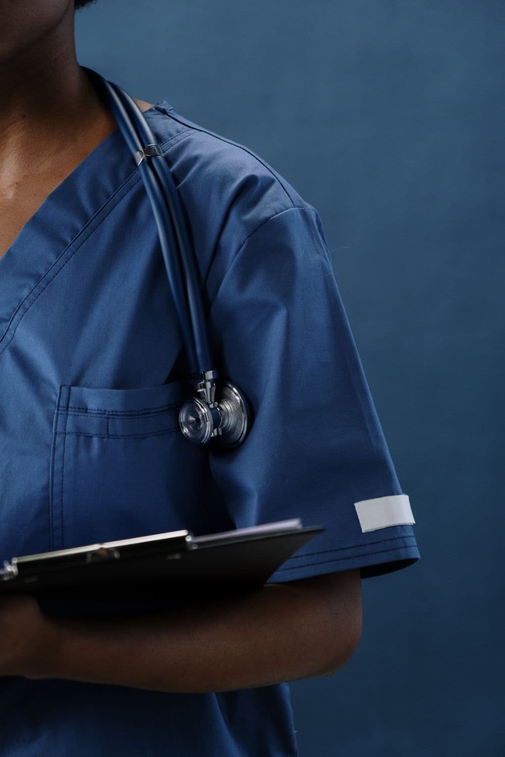 A nurse in navy scrubs holding a clipboard, stethoscope around the neck