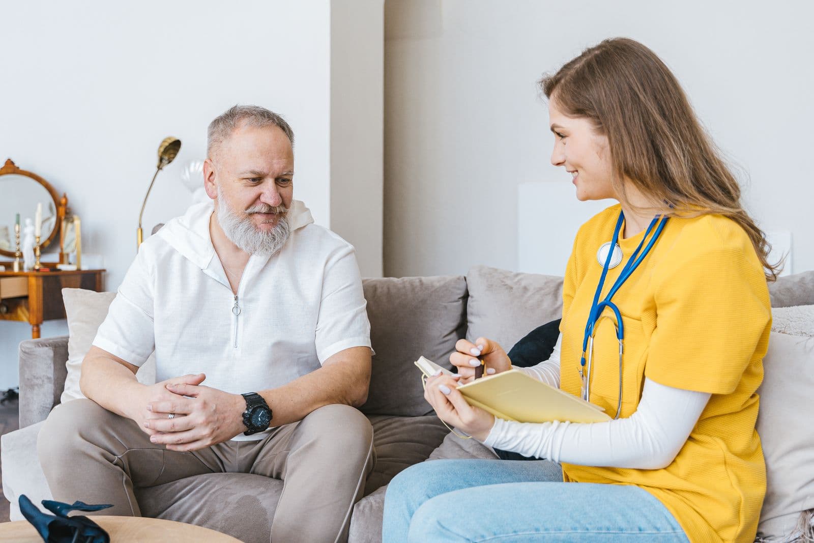 A caregiver in a yellow uniform with a clipboard chatting with a smiling older man on his sofa