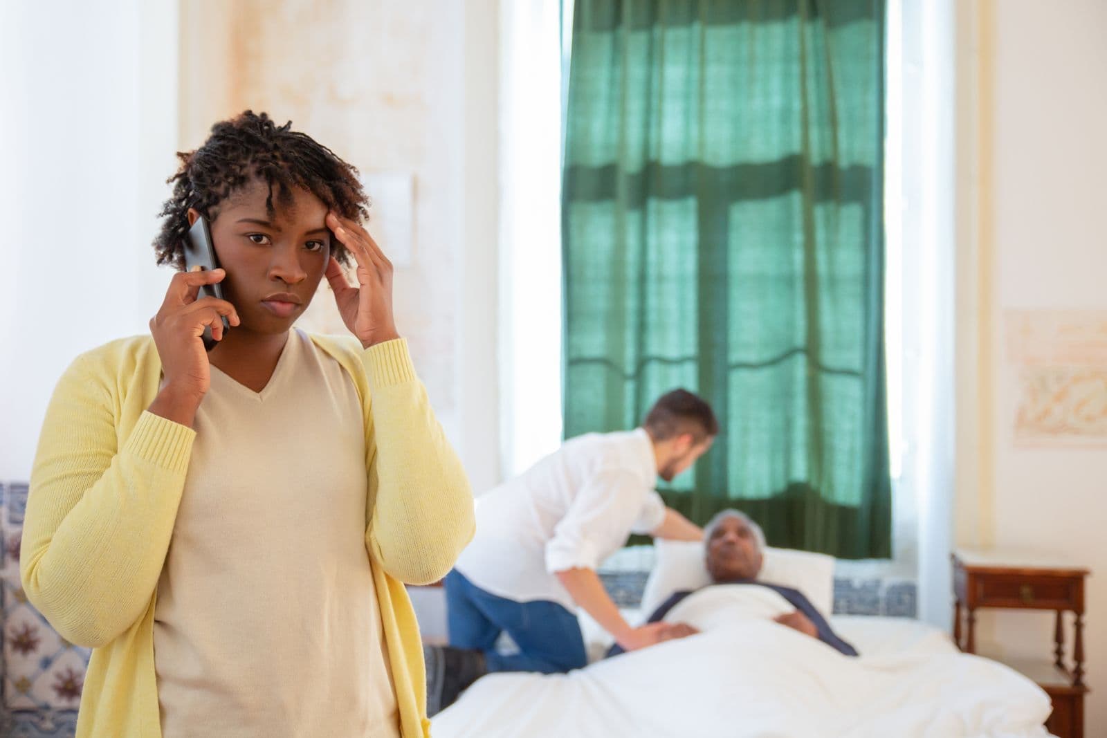 A worried woman on the phone while a caregiver settles an elderly man into bed behind her