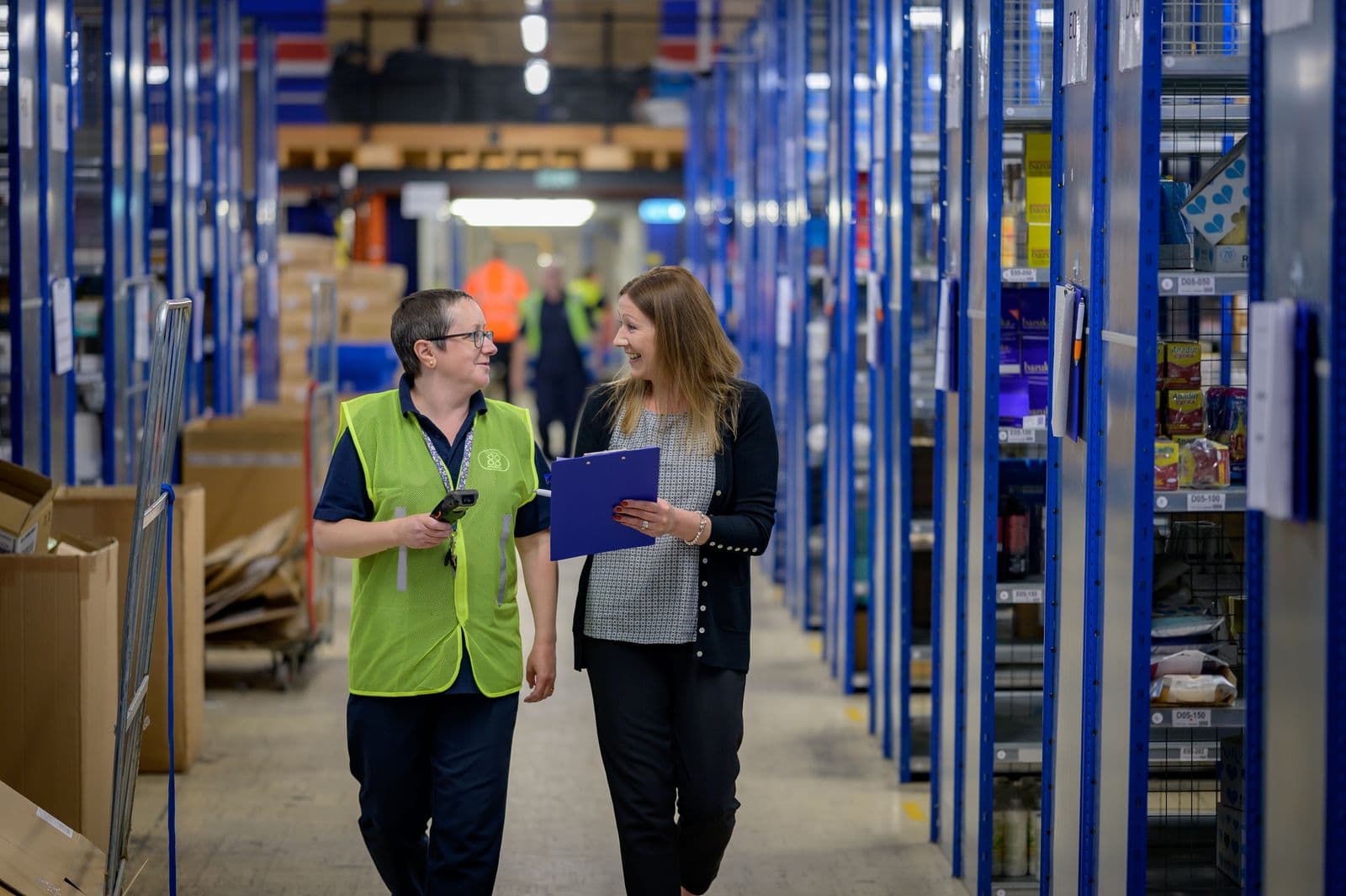 A supervisor in a hi-vis vest and a colleague with a clipboard talking as they walk down a warehouse aisle