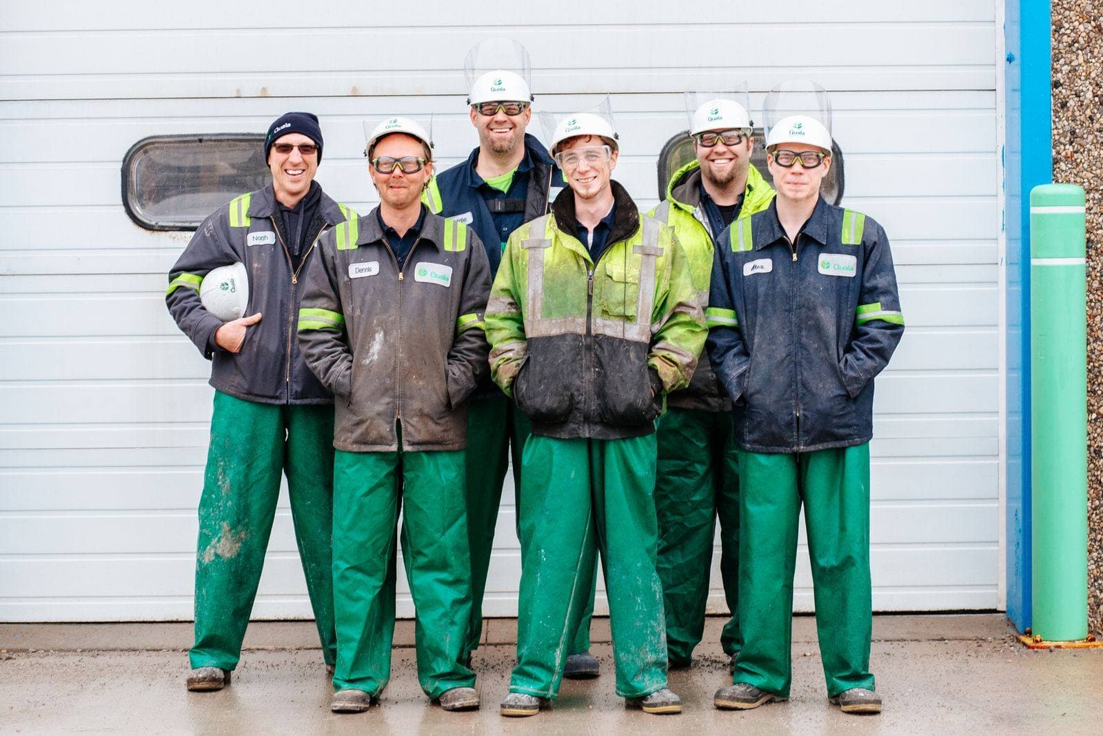 A crew of six industrial workers in hard hats and work jackets standing together