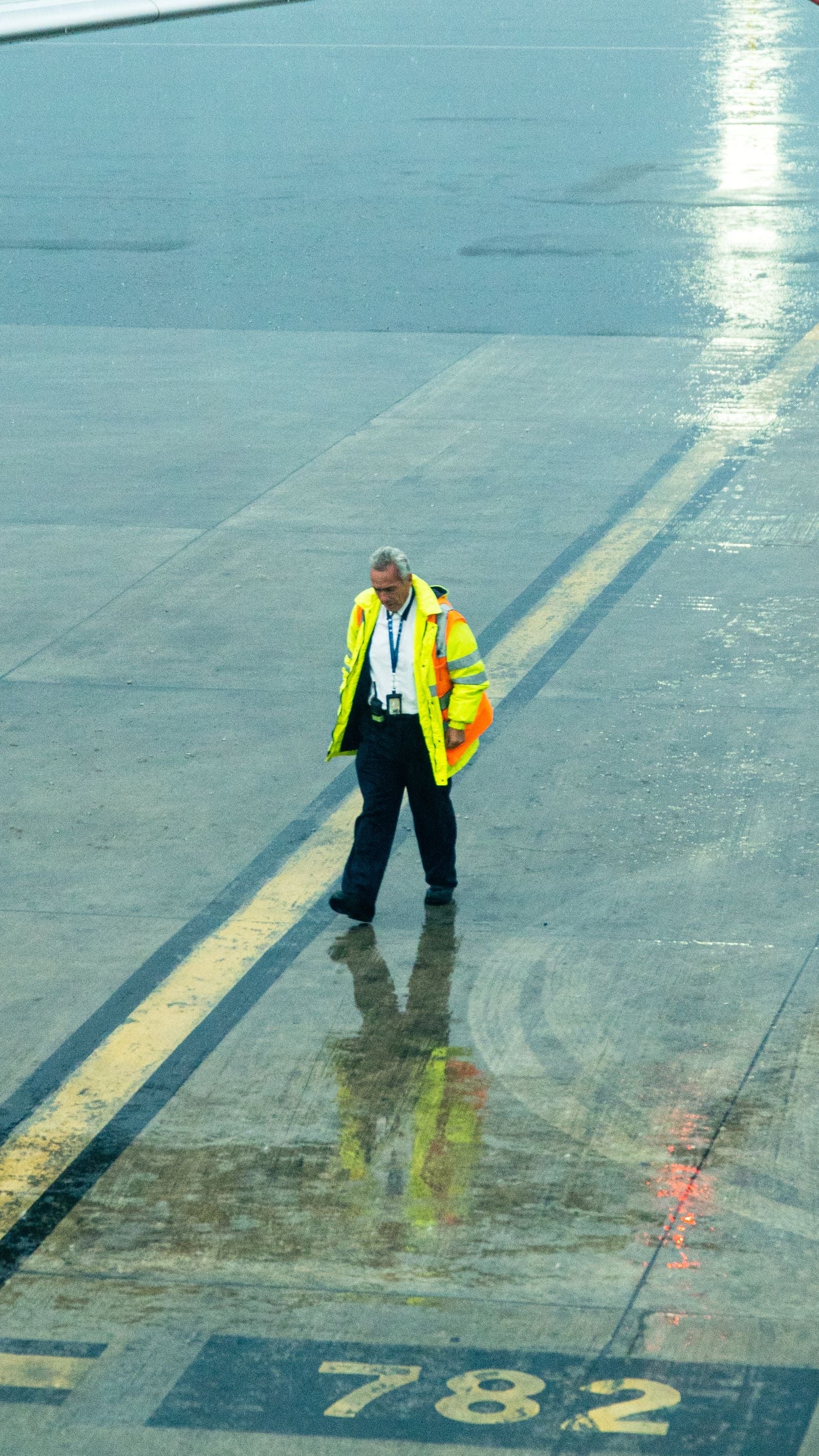 A ground handler in a hi-vis jacket walks across a wet apron beside the yellow stand markings