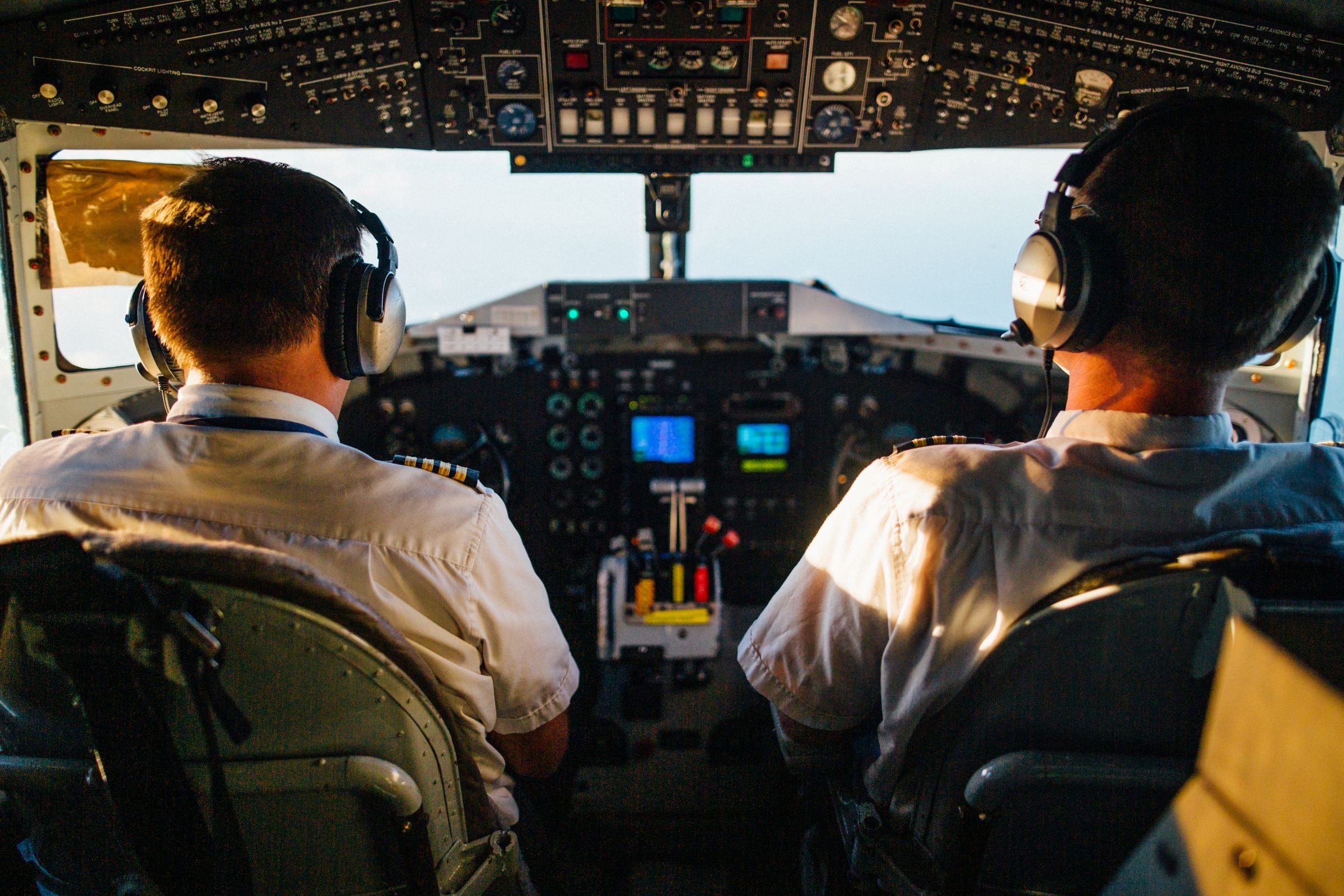 Two pilots in headsets and white shirts fly an aircraft, seen from the back of the flight deck