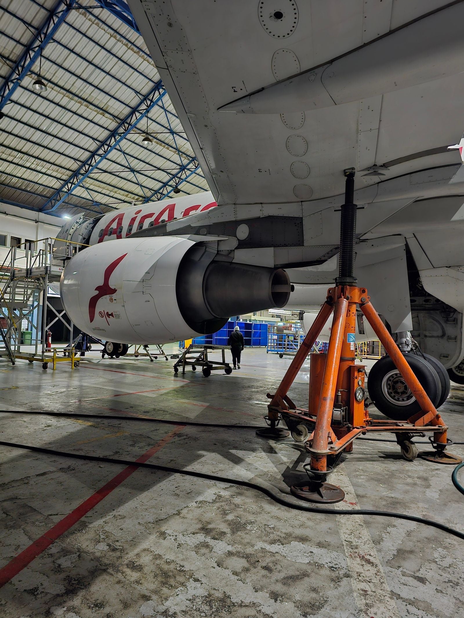 An airliner stands on an orange jack in a maintenance hangar, its engine cowling beside the main landing gear