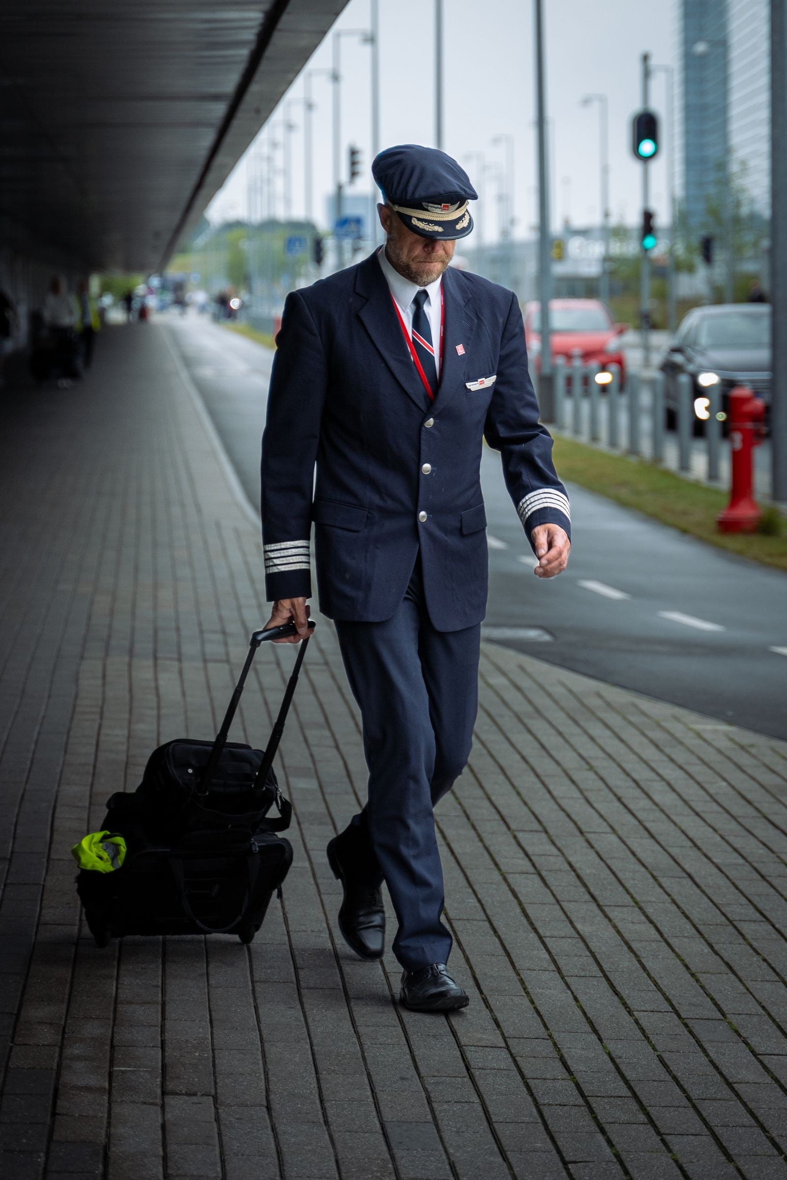 A pilot in a dark uniform and cap pulls a flight bag along a pavement outside an airport terminal