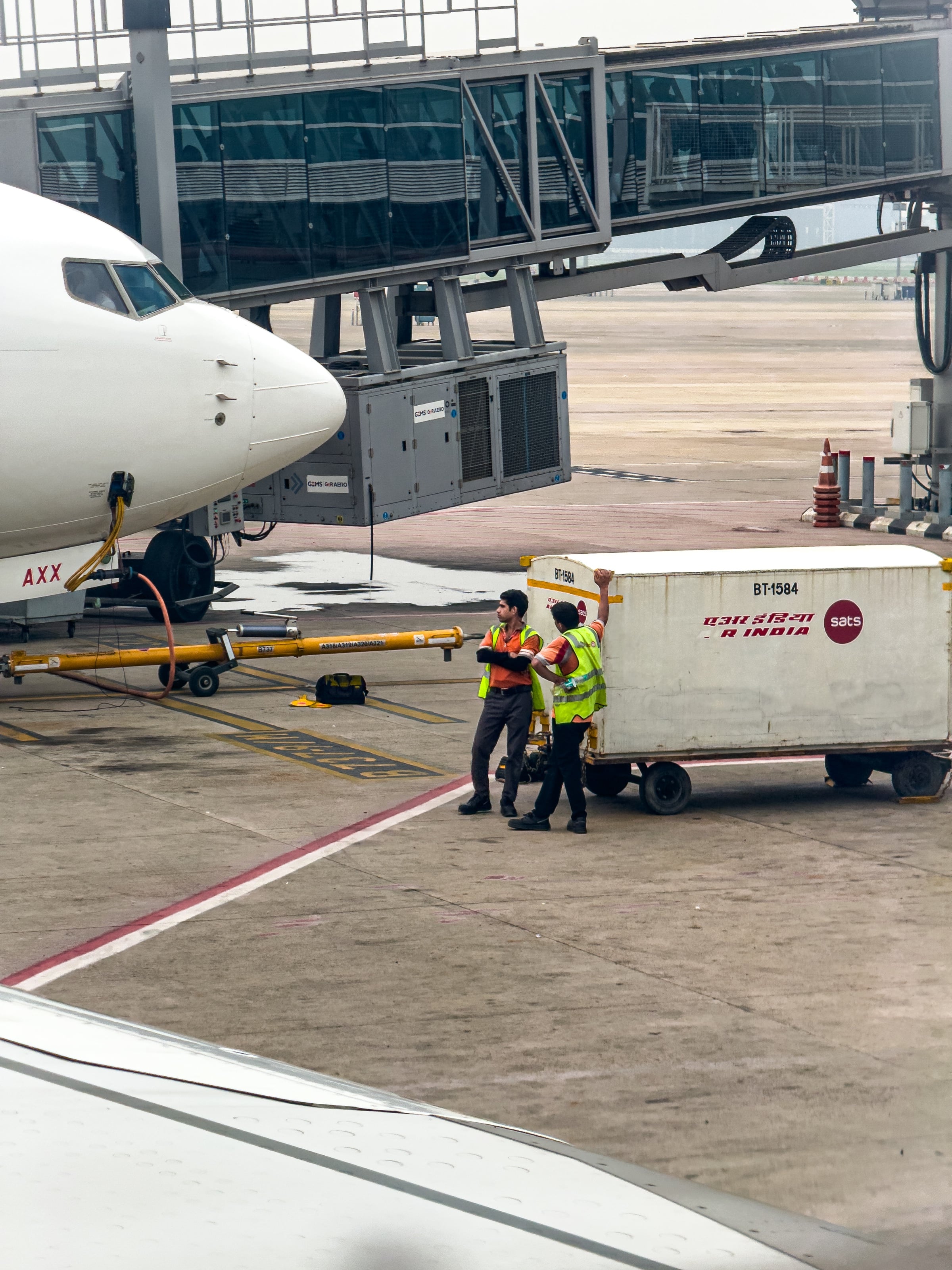 Two ground handlers in hi-vis vests talk beside a baggage container at the nose of an airliner on stand