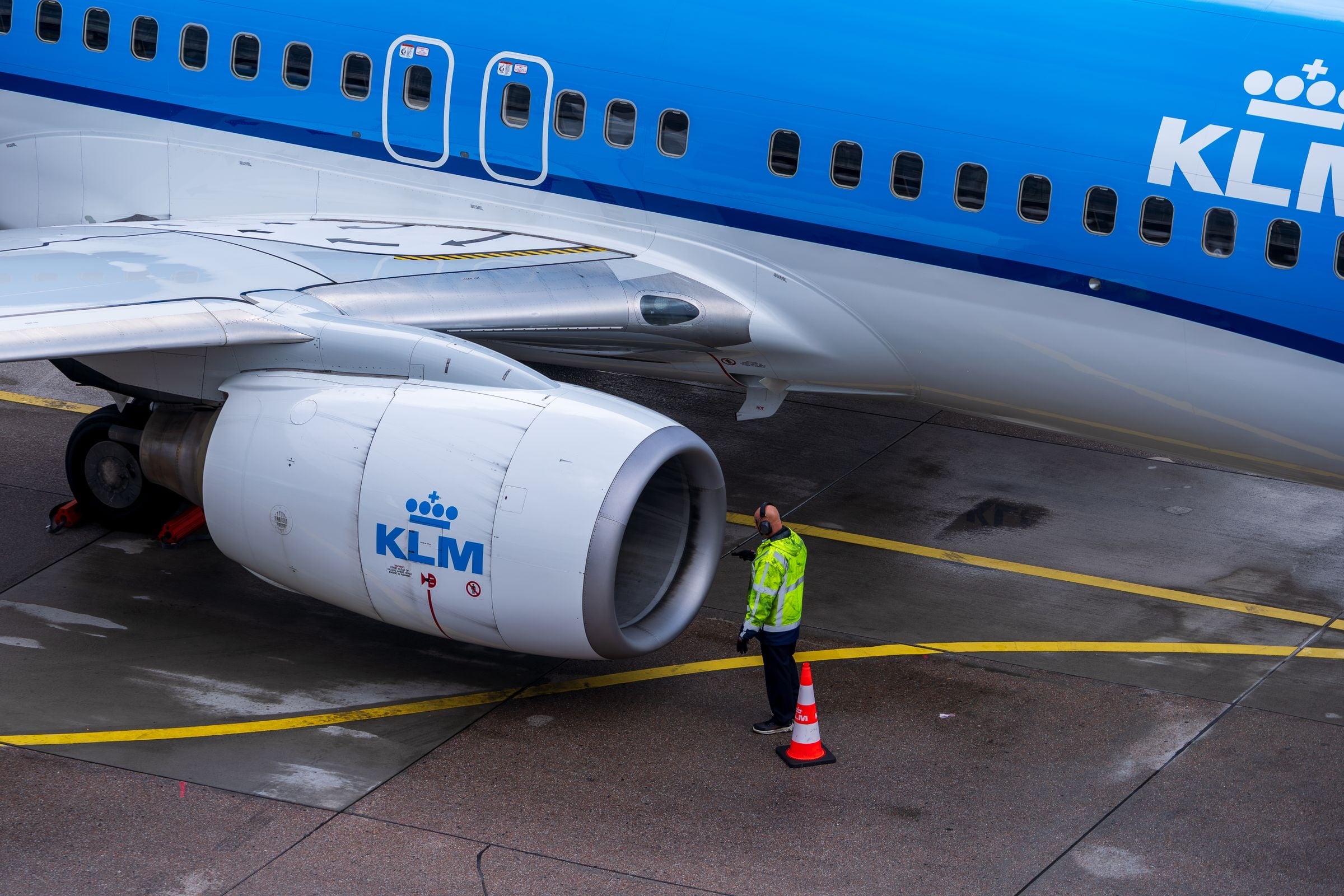 An engineer in a hi-vis jacket stands by a cone beside the engine of a parked KLM airliner