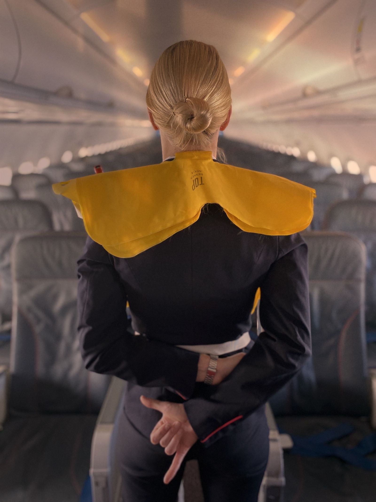 A cabin crew member, seen from behind, wears a yellow life jacket for a safety demonstration in an aircraft aisle
