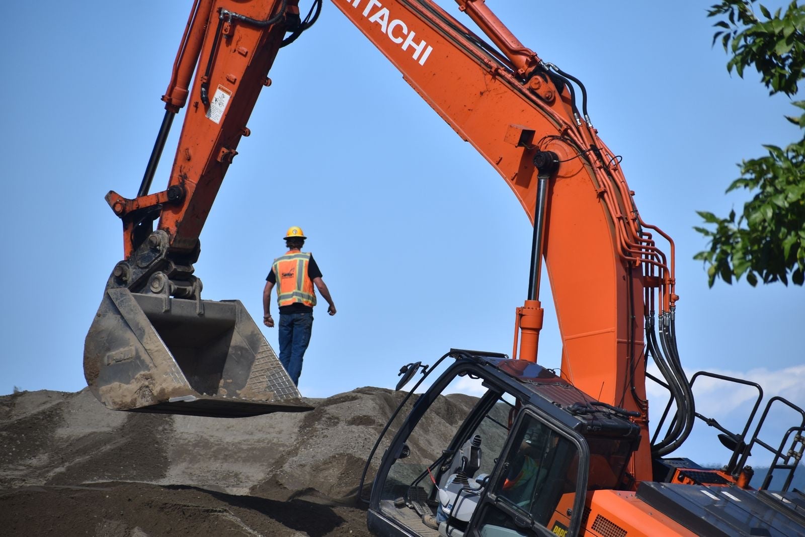 An operative in hi-vis walking on a spoil heap beneath the raised arm of an orange excavator
