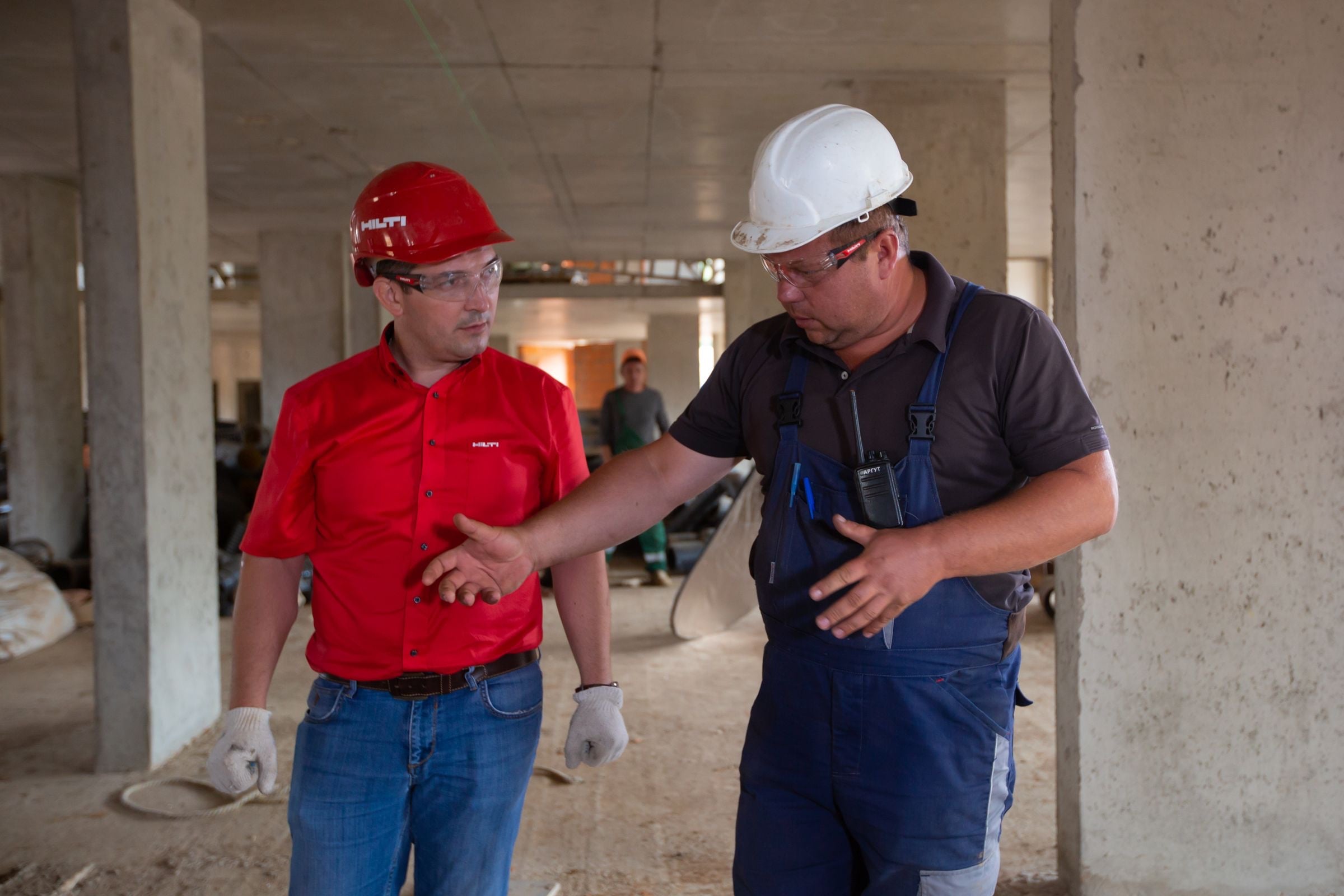 A site manager in a red hard hat talks with an operative in a white hard hat inside a concrete-framed building