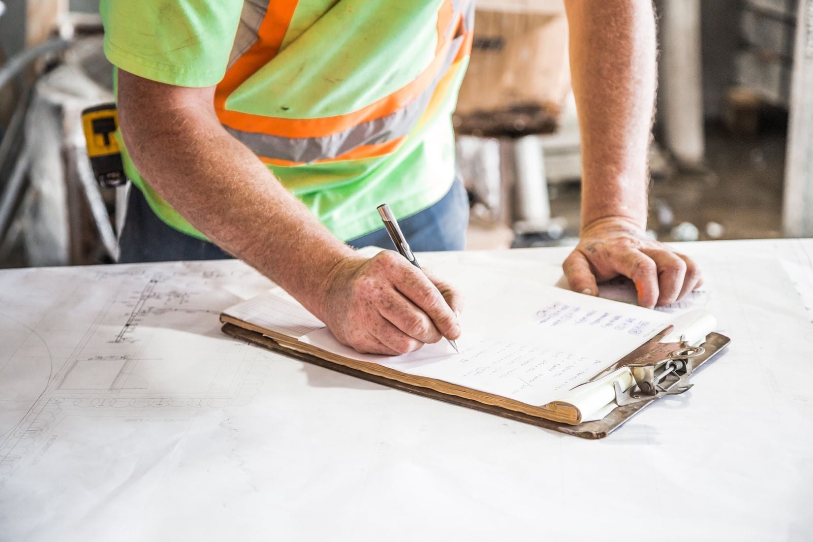 A worker in a hi-vis vest filling in a form on a clipboard laid on drawings