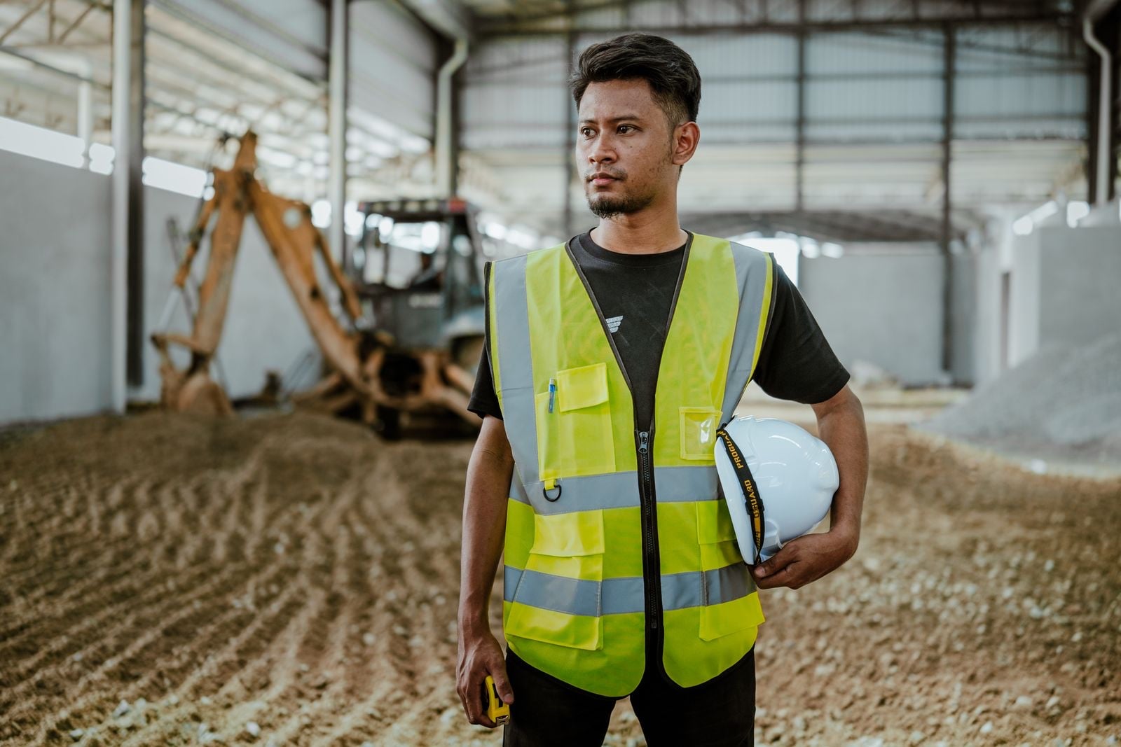 An operative in a yellow hi-vis vest holding a white hard hat, with a digger in the shed behind him