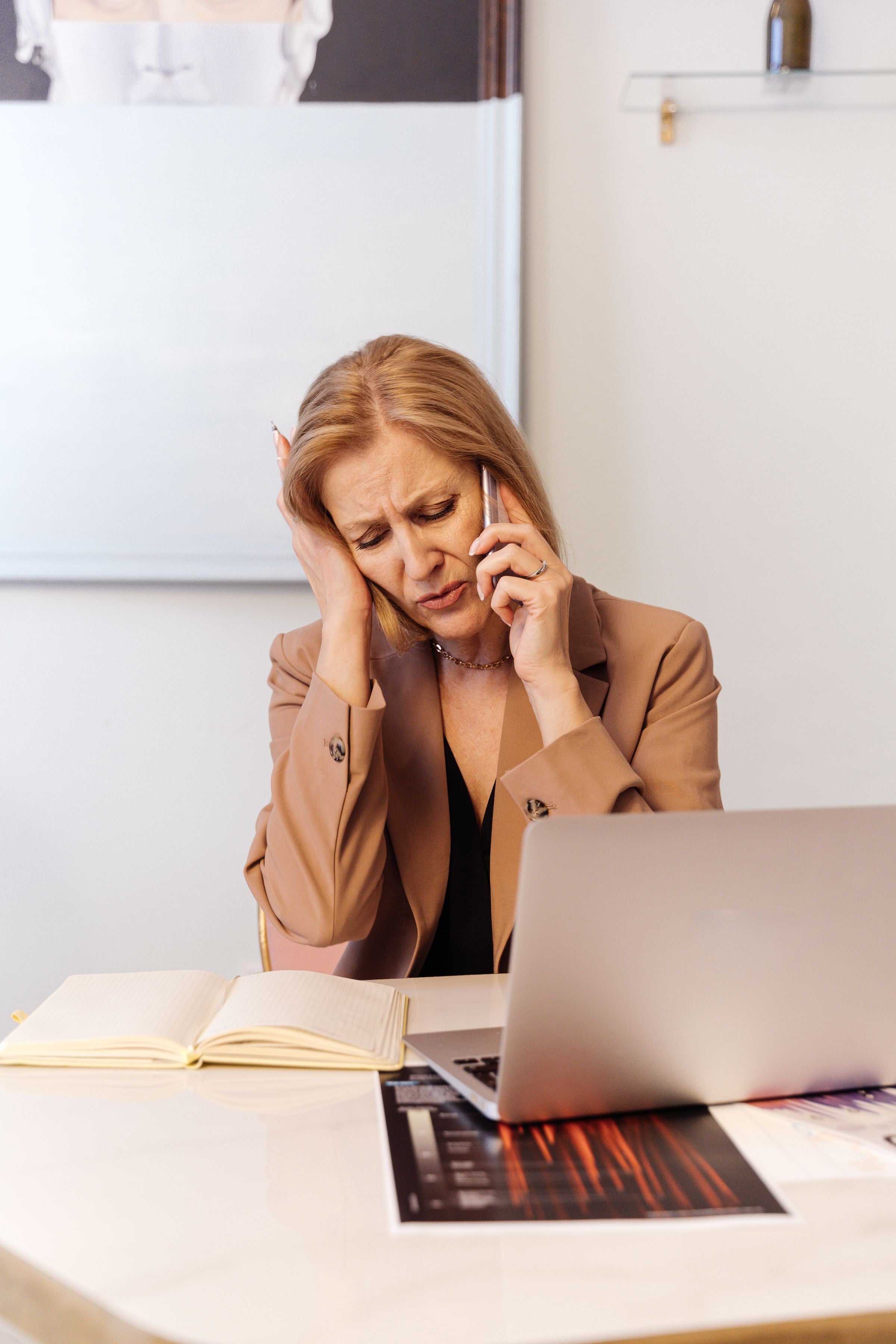 A worried woman on her mobile at a desk with an open laptop and notebook