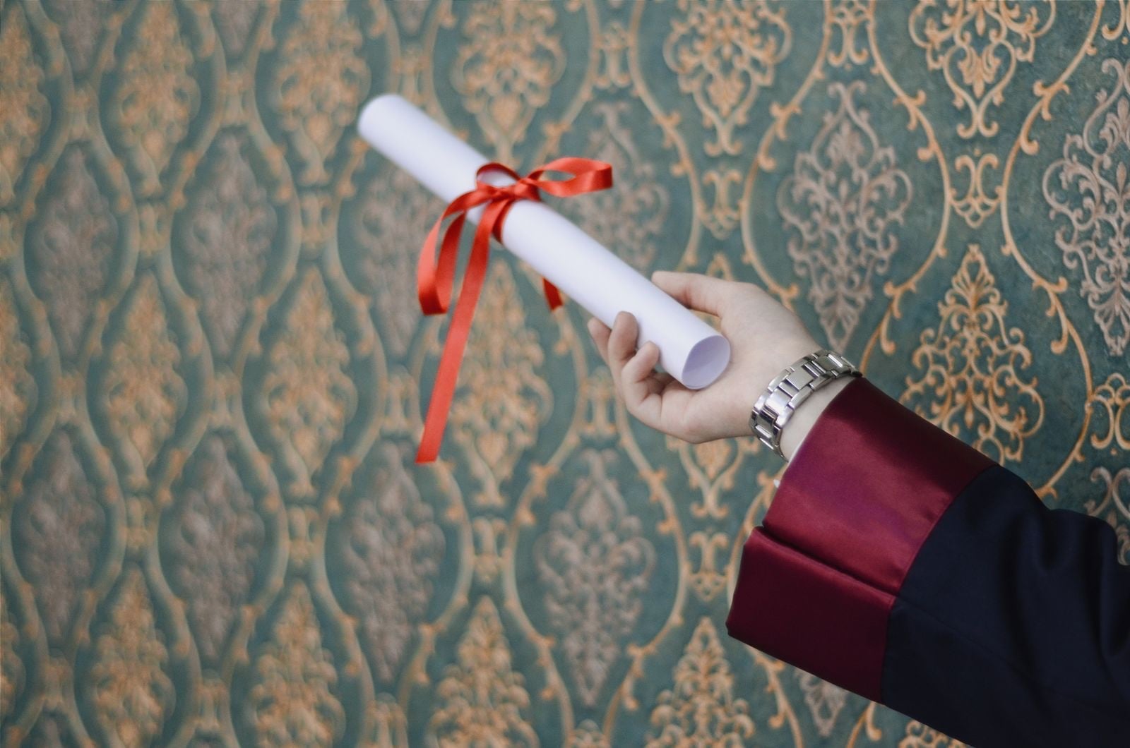 A hand in a graduation gown sleeve holding a rolled certificate tied with a red ribbon