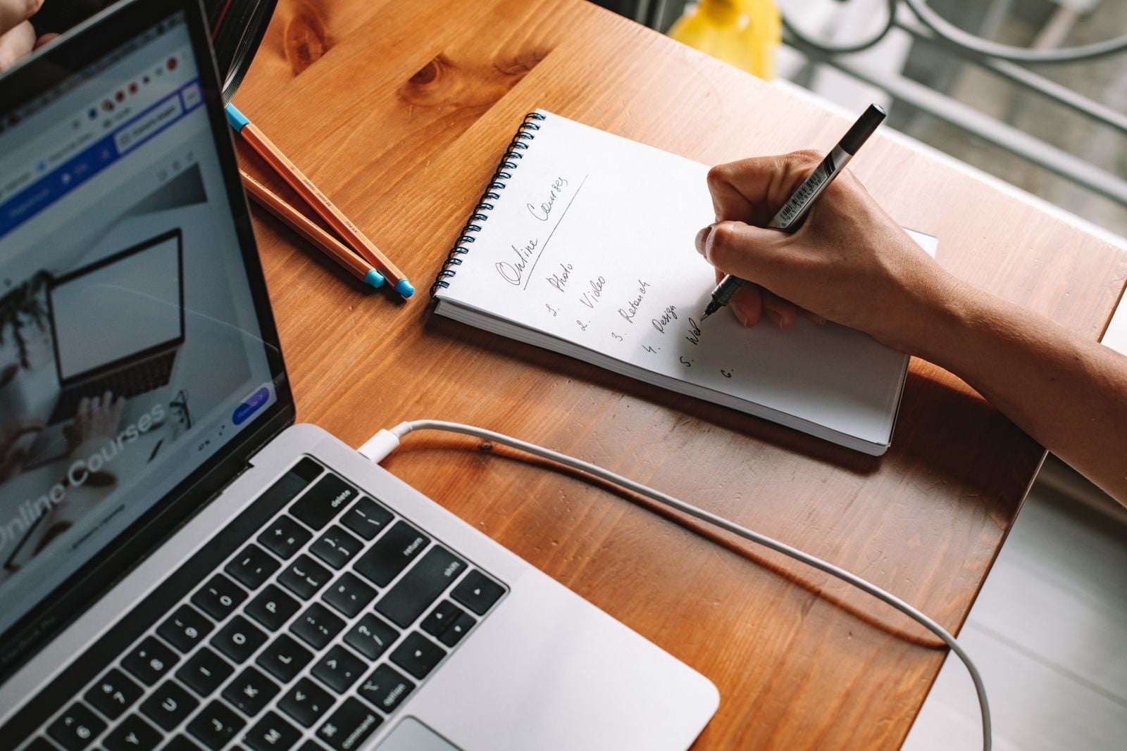 A person writing a list in a notepad beside an open laptop on a wooden desk