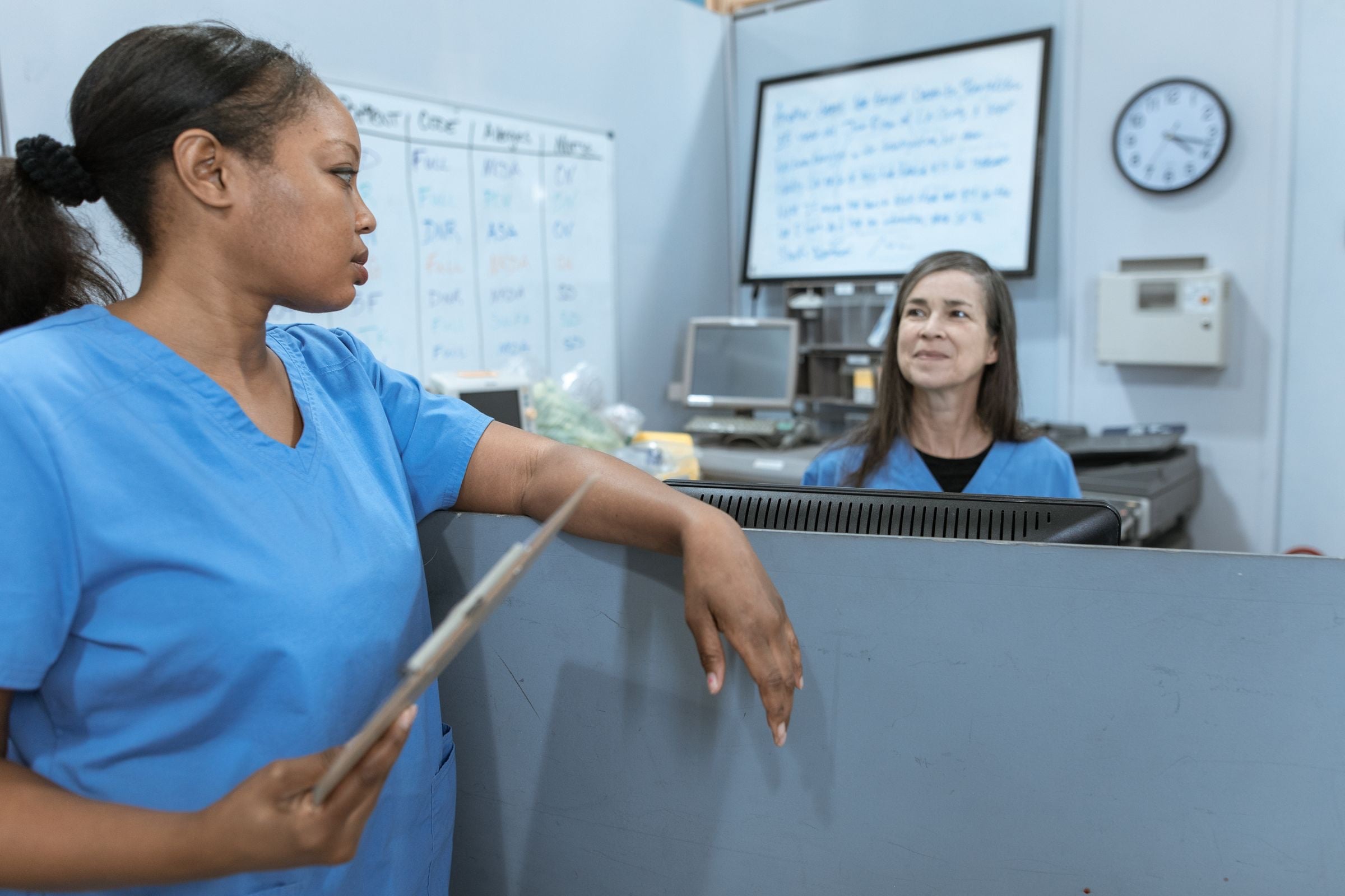 A nurse in blue scrubs talking with a colleague at a ward nurses’ station