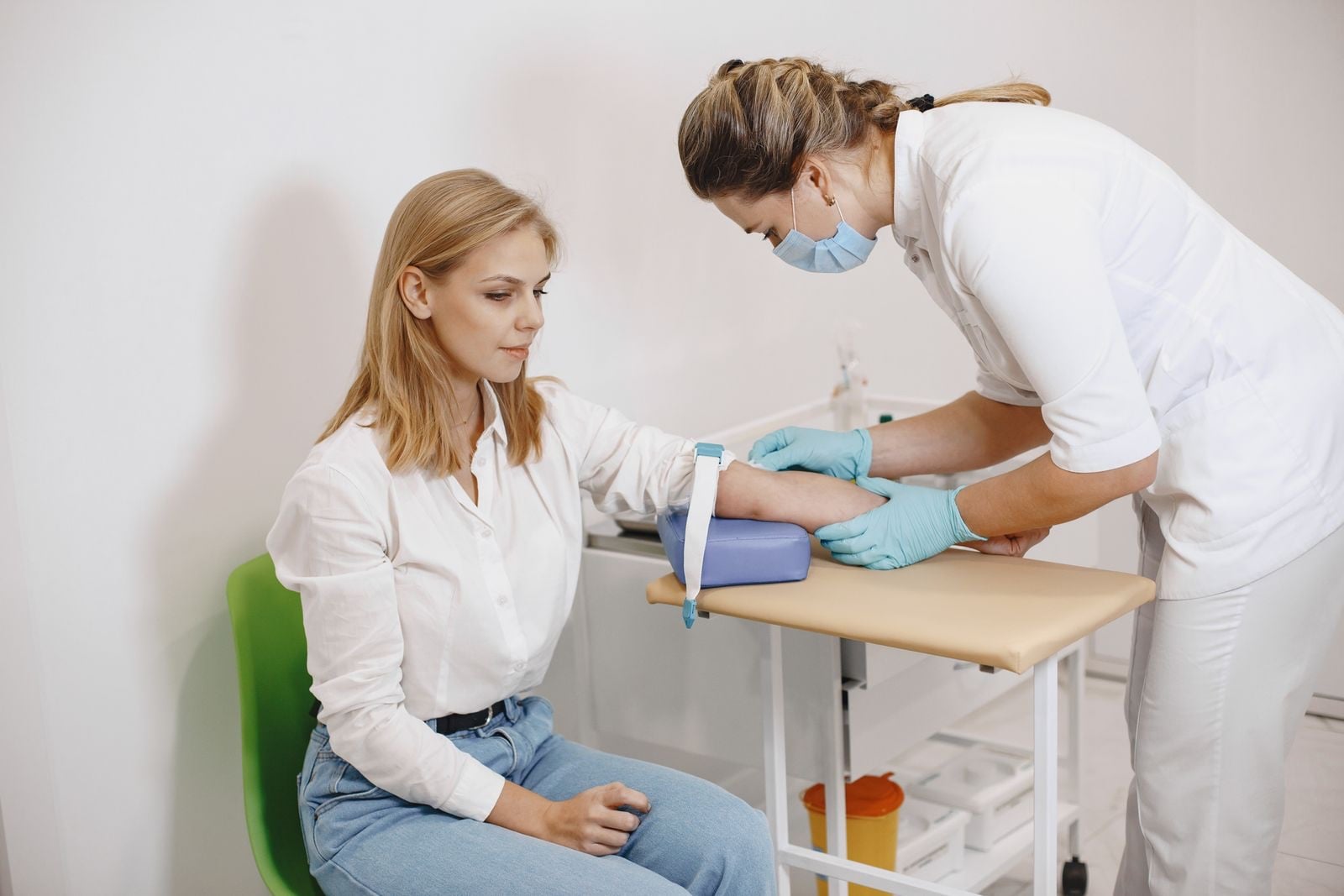 A nurse in a white tunic taking a blood sample from a seated woman’s arm
