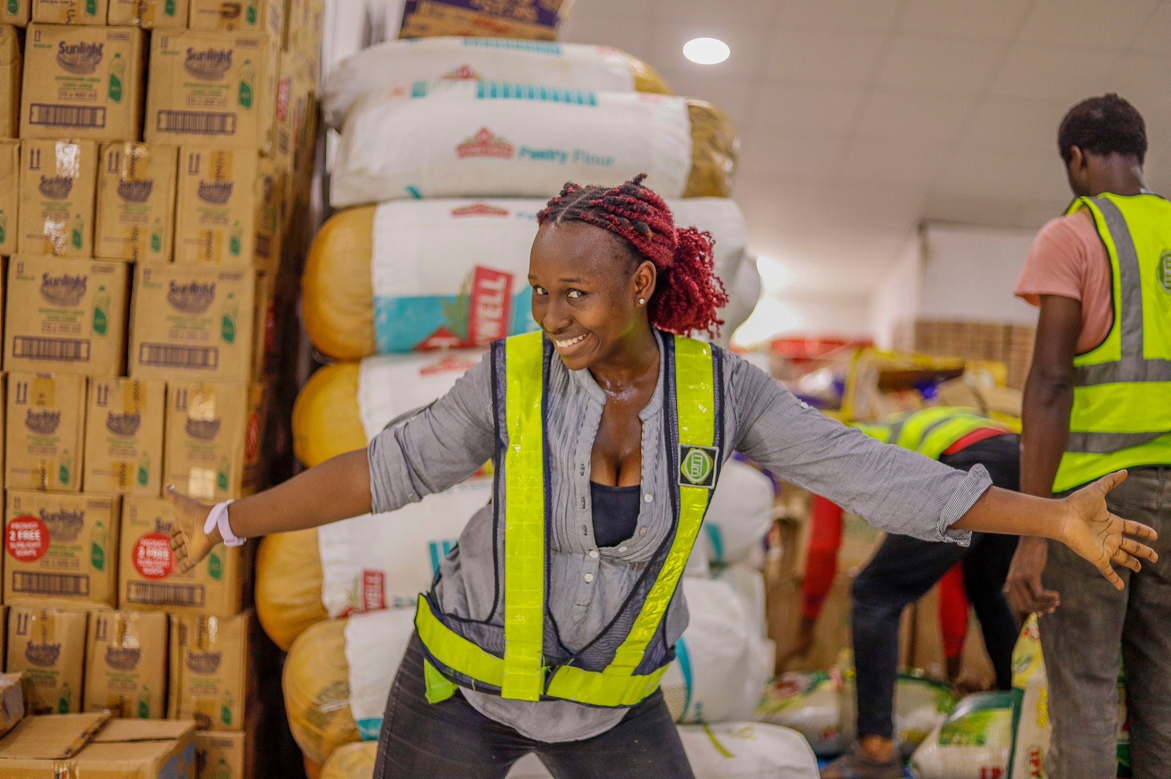 A smiling worker in a hi-vis vest with her arms out among sacks and boxes in a busy warehouse, a colleague behind her