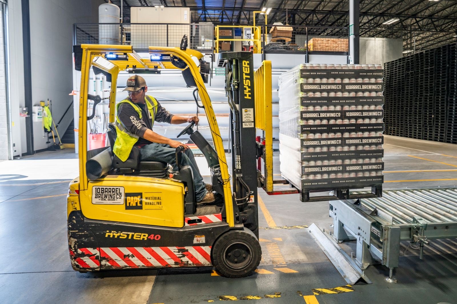A forklift operator in a hi-vis vest moving a loaded pallet past a conveyor in a warehouse