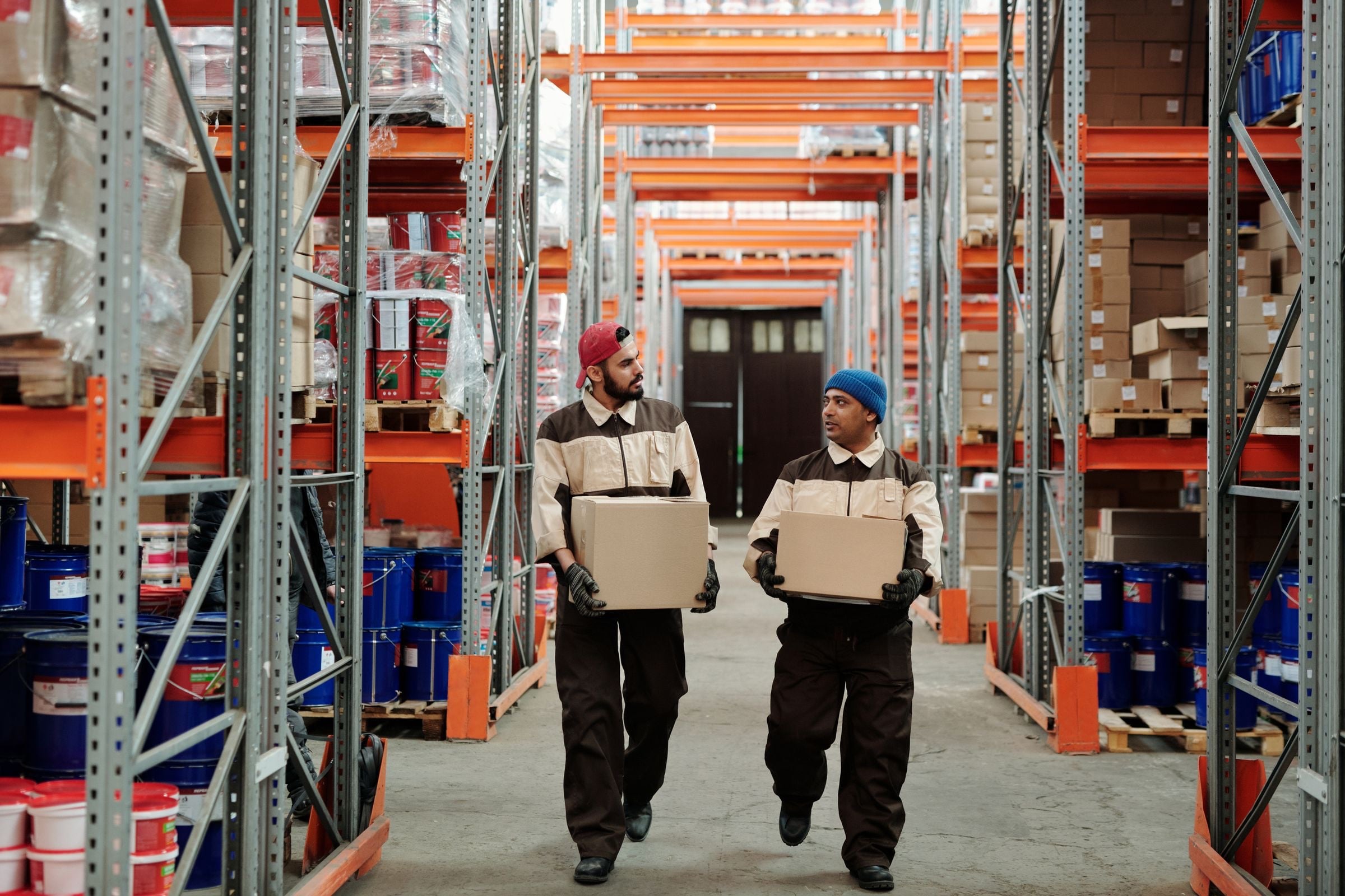 Two warehouse workers in work jackets carrying boxes down an aisle between orange pallet racking