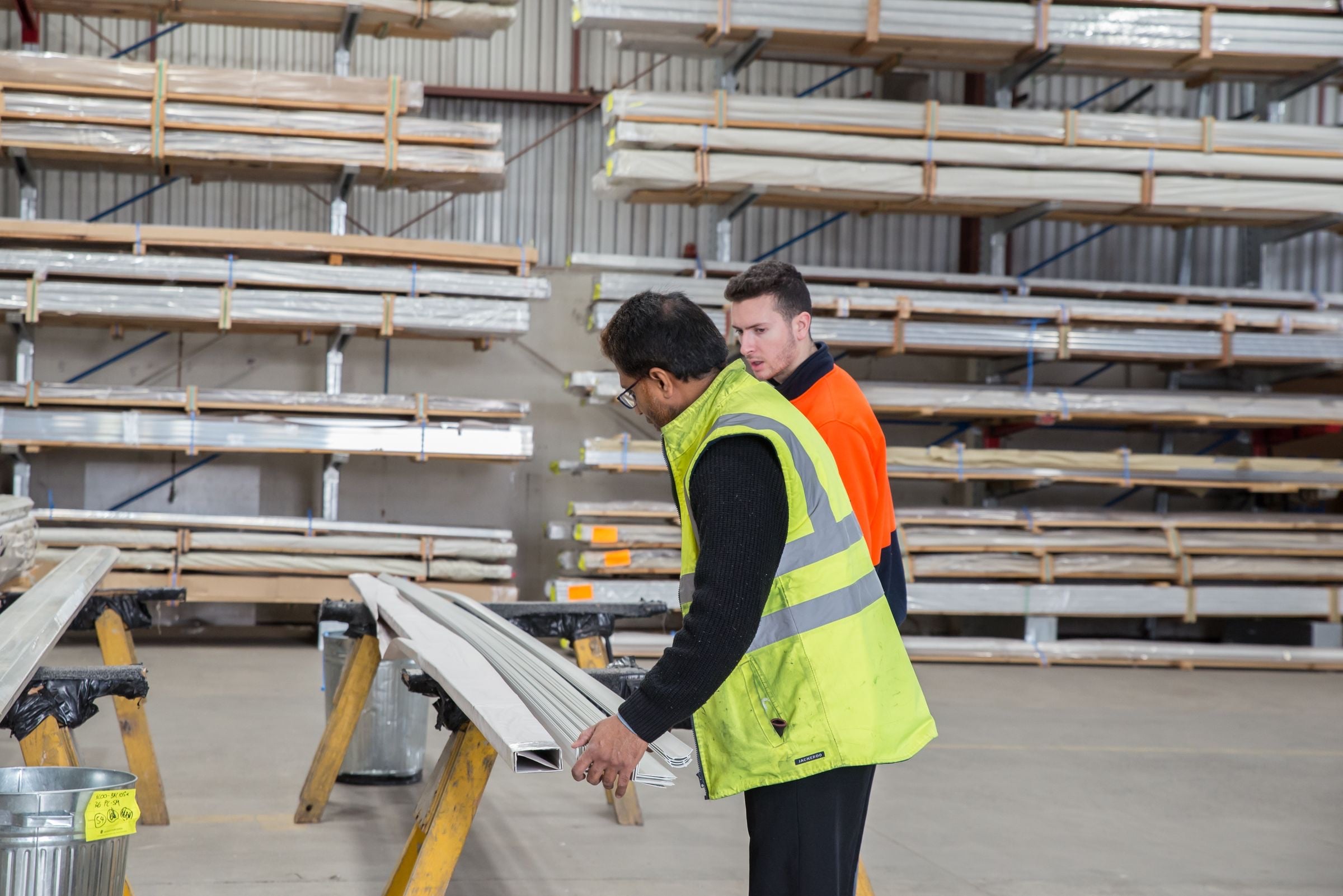 Two workers in hi-vis vests checking lengths of material on trestles in a racked warehouse