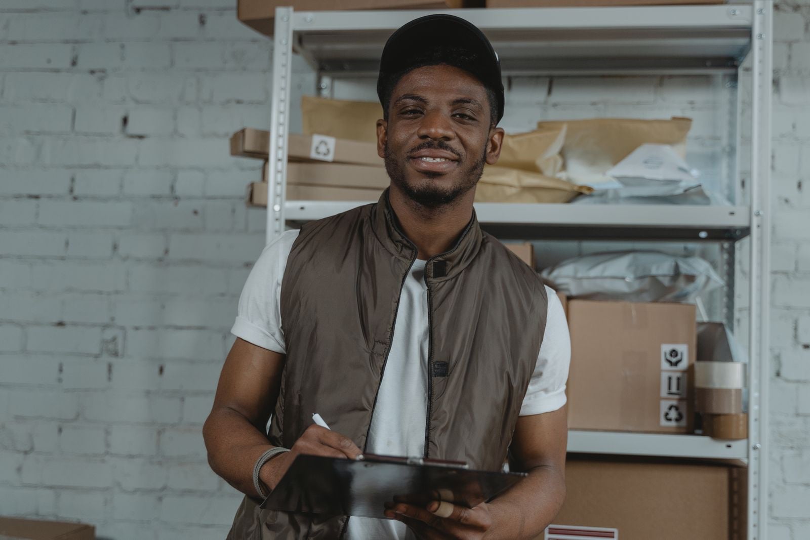 A smiling warehouse worker in a cap and gilet filling in a form on a clipboard in a stockroom